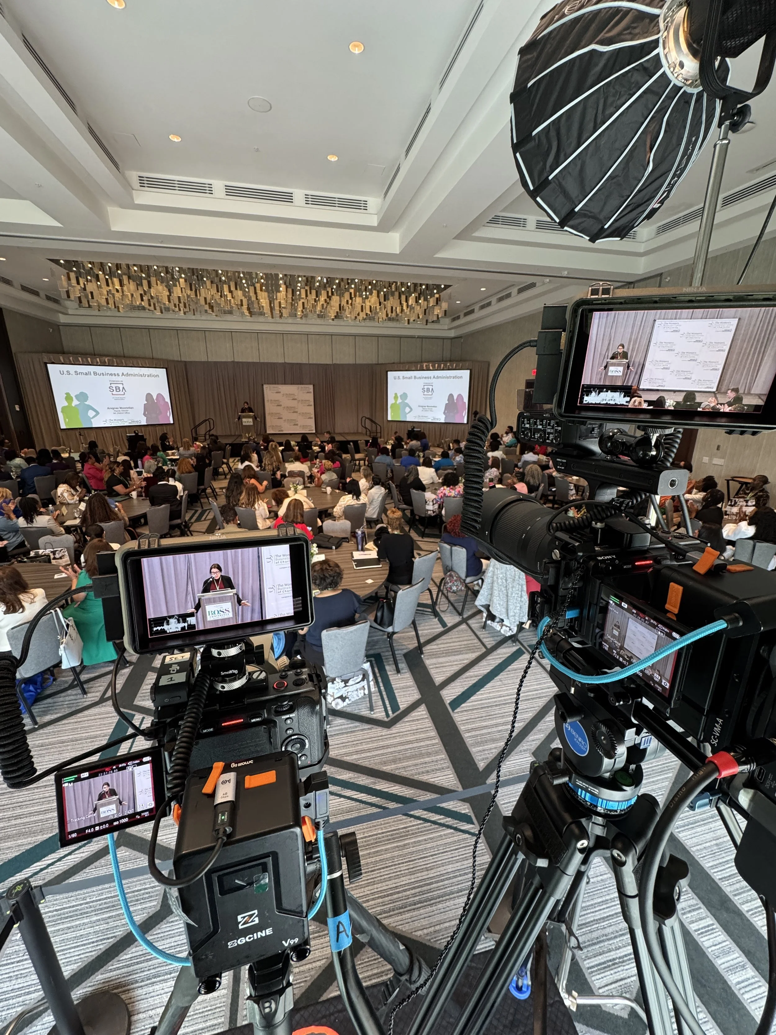 Camera setup recording a speakers at a conference stage with audience present in a hotel ballroom.