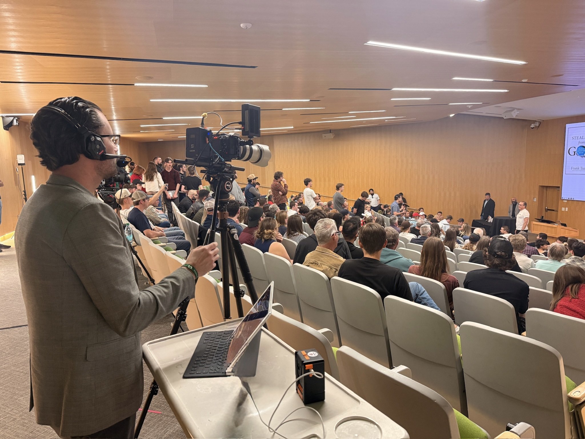 A man operating a professional video camera on a tripod records at an indoor conference with an audience. The audience is seated facing a stage where a panel of speakers is present, and a large screen displays a presentation.
