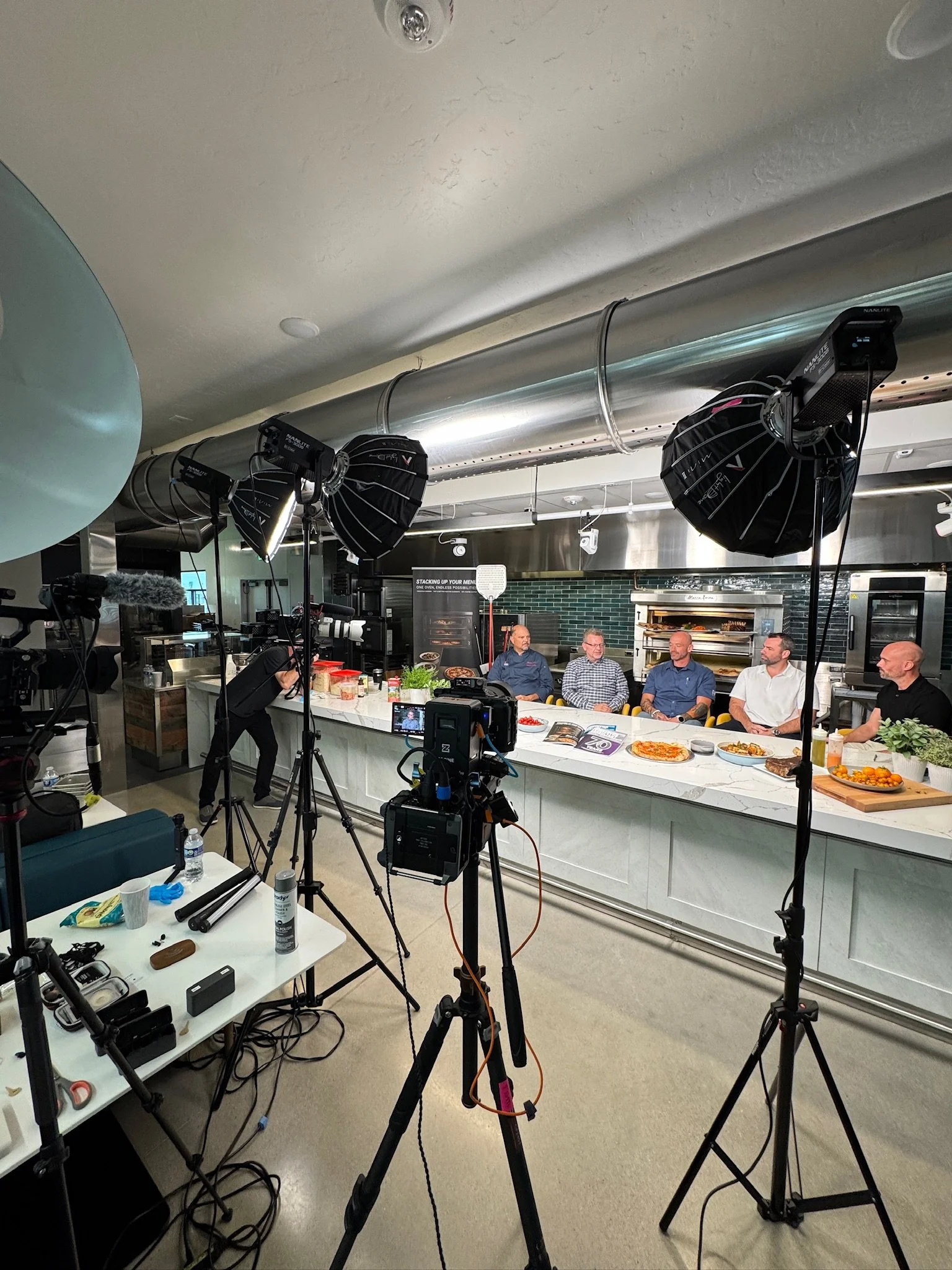 A professional video shoot in a modern kitchen studio with five men sitting behind a white counter, surrounded by lighting equipment and cameras facing them.