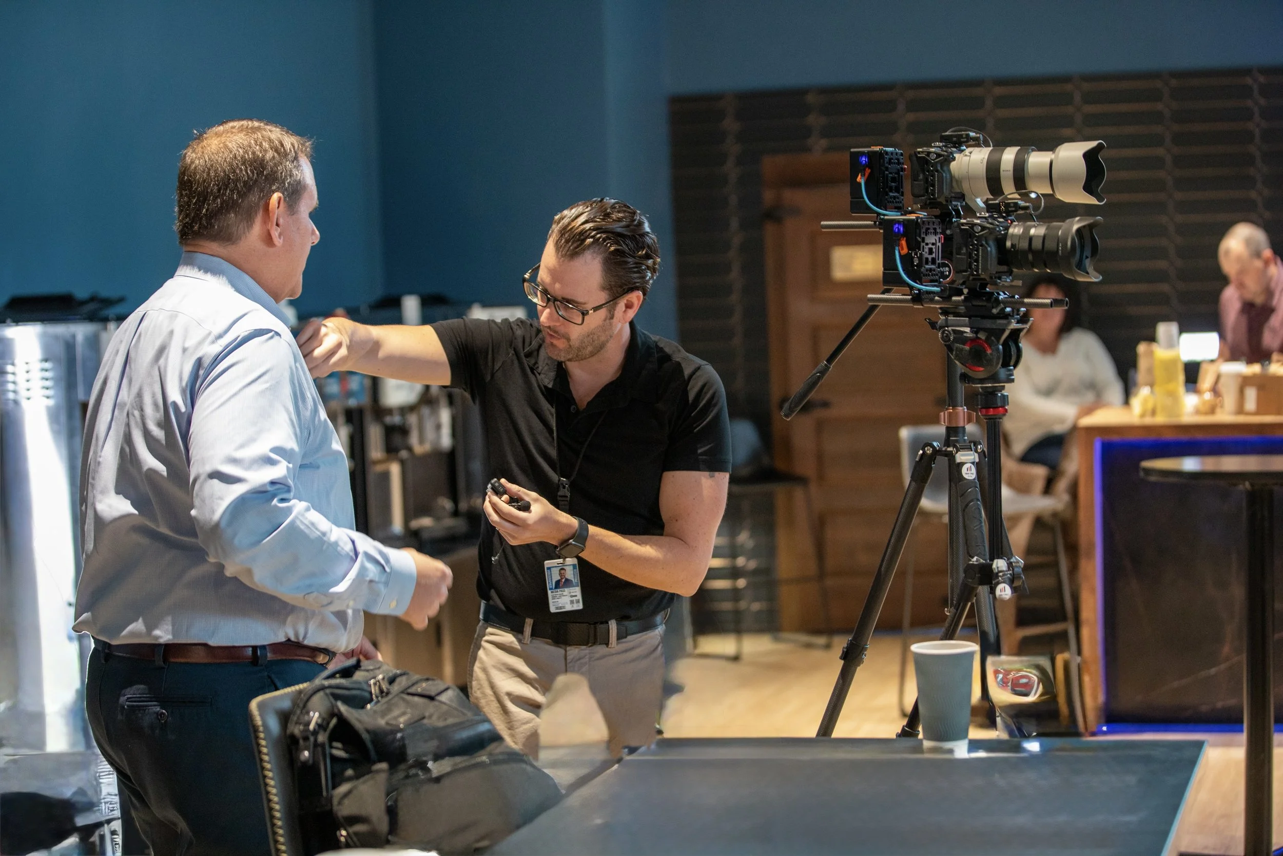 A man with a badge and a black shirt adjusts the shoulder of another man in a light blue shirt in a room with cameras and people working in the background.
