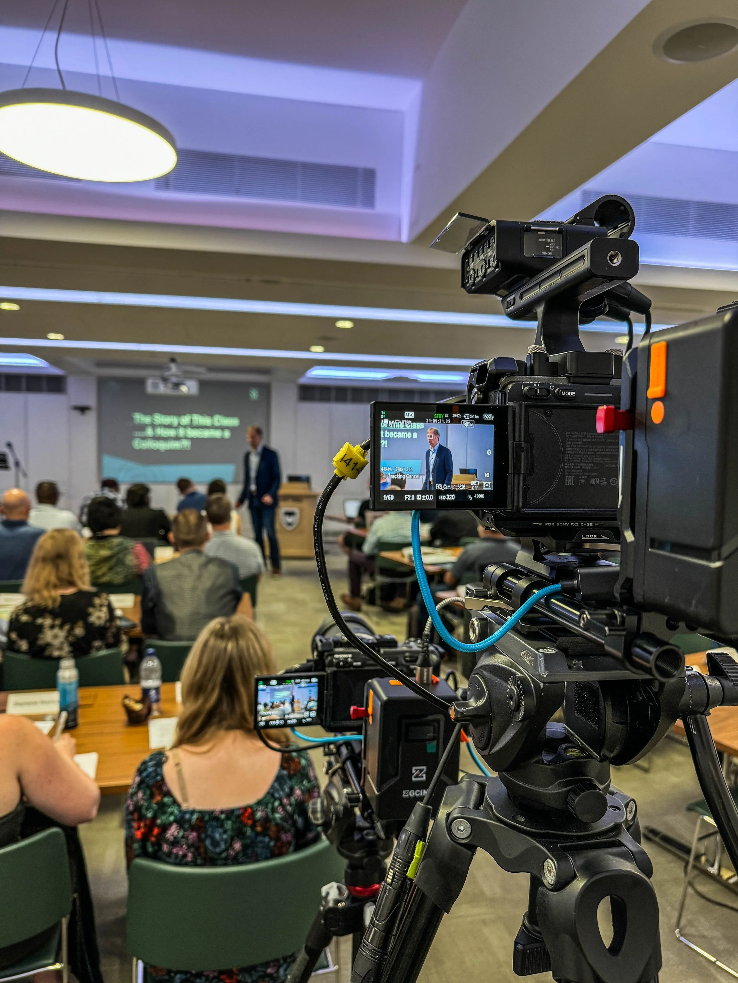 Camera filming a speaker at a conference or lecture in a classroom or conference room with an audience.