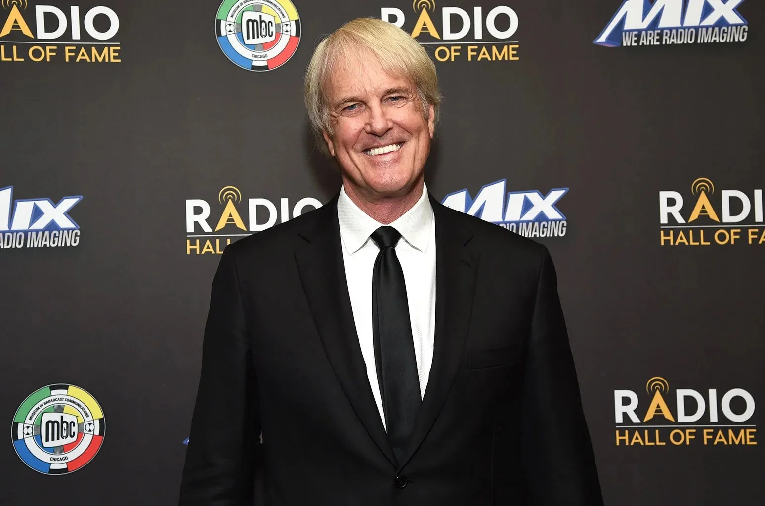 A man in a black suit and tie smiling at a Radio Hall of Fame event with a backdrop featuring various logos, including Radio Hall of Fame, MBC Chicago, and MIX Radio Imaging.