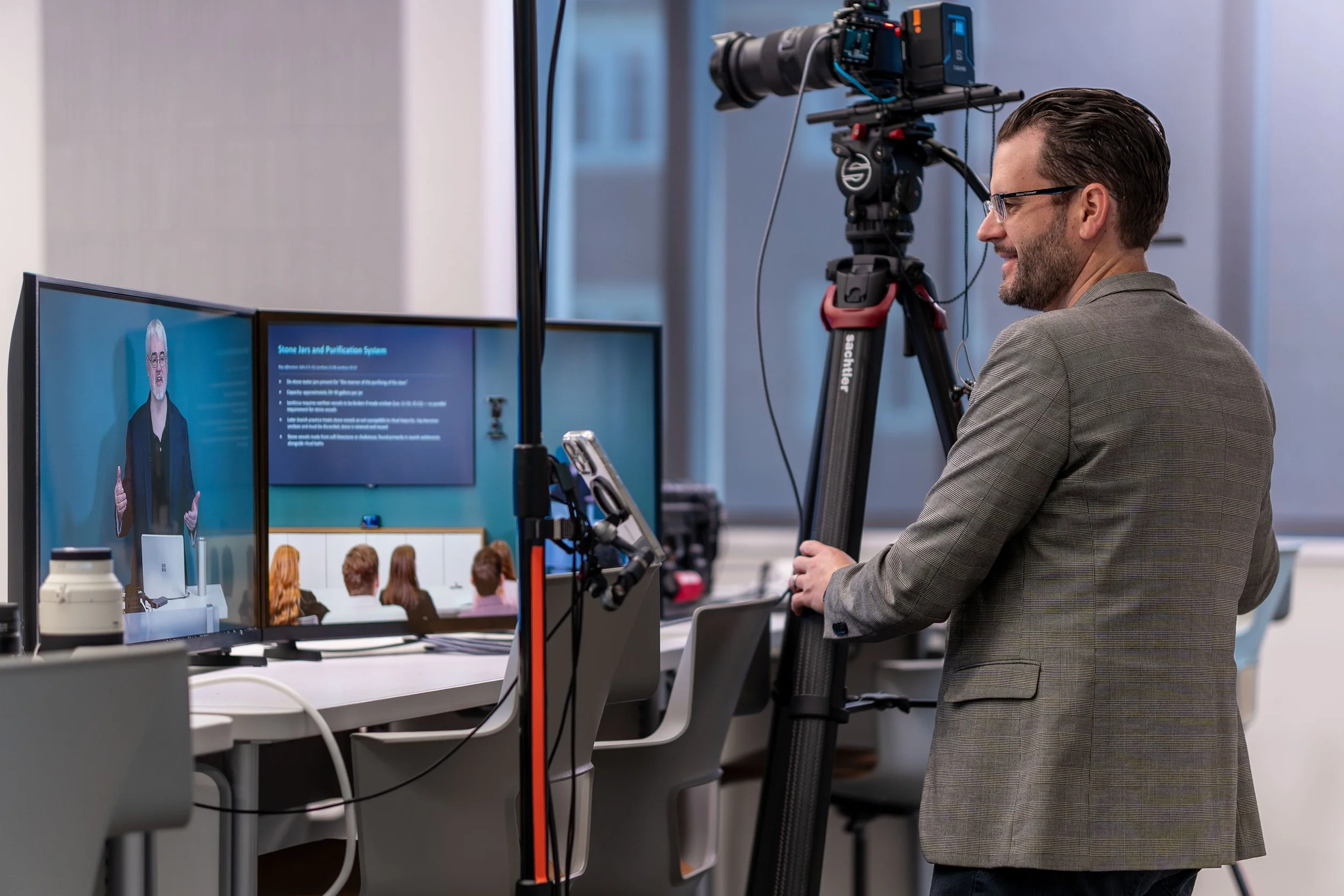 A man with glasses and a checkered blazer standing in front of a camera setup, filming a presentation with two monitors.