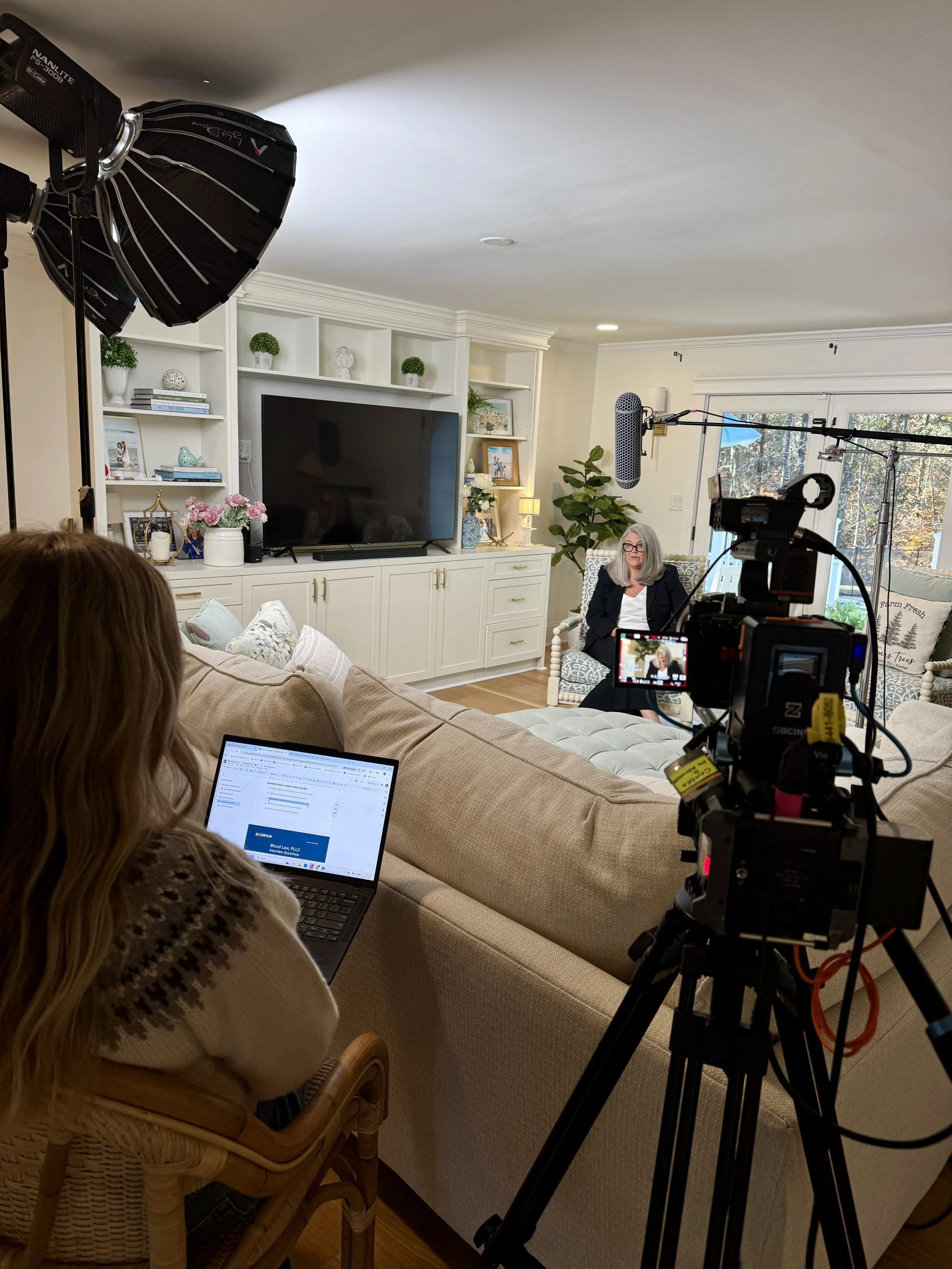 A woman is being recorded in a home studio setup on a beige sofa, with a camera on a tripod and a microphone suspended above her. A crew member is working on a laptop, adjusting settings for a video shoot being conducted in a living room with white cabinetry, a large TV, and decor.
