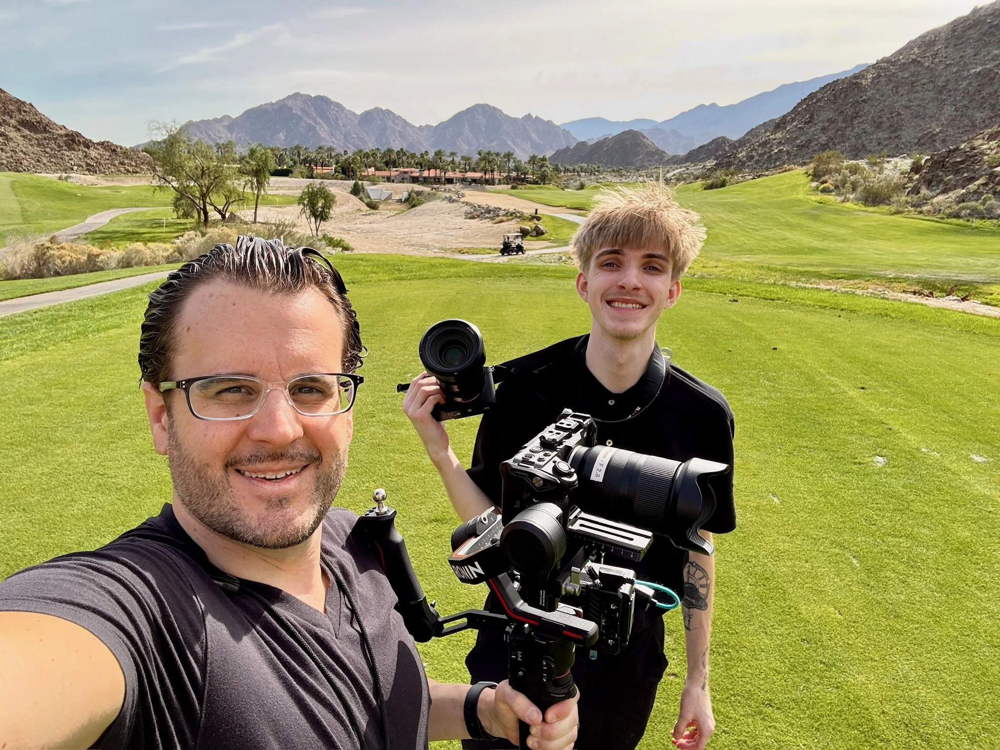 Two men on a golf course field. One man is taking a selfie while the other is holding a professional camera with a large lens. The background shows mountains, trees, and a golf course landscape.