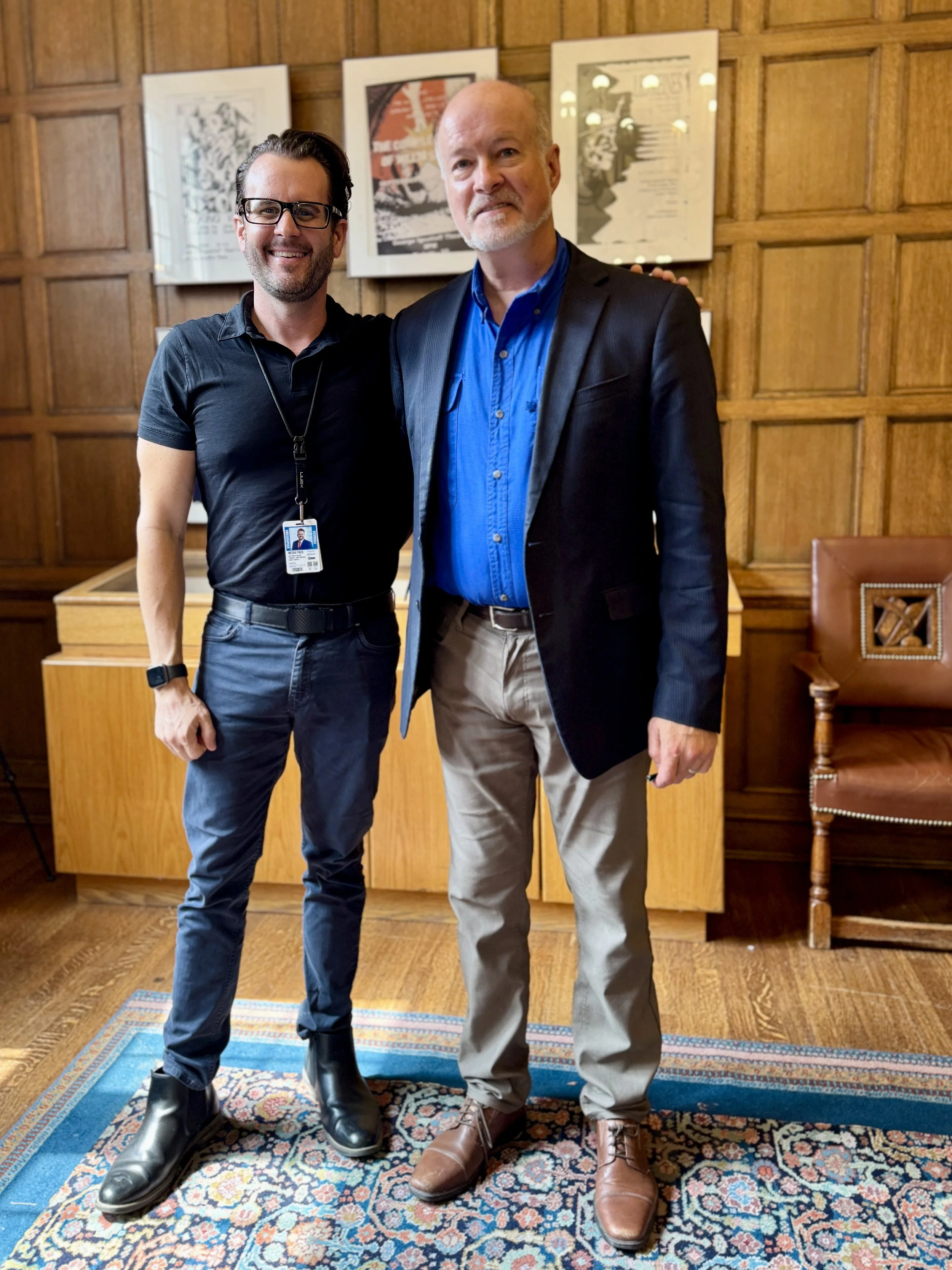 Two men standing next to each other in a room with wooden paneling and framed art on the wall. The man on the left is smiling, wearing glasses, a black polo shirt, and dark pants. The man on the right is wearing a blue shirt, a black blazer, and light-colored pants. Both are smiling.