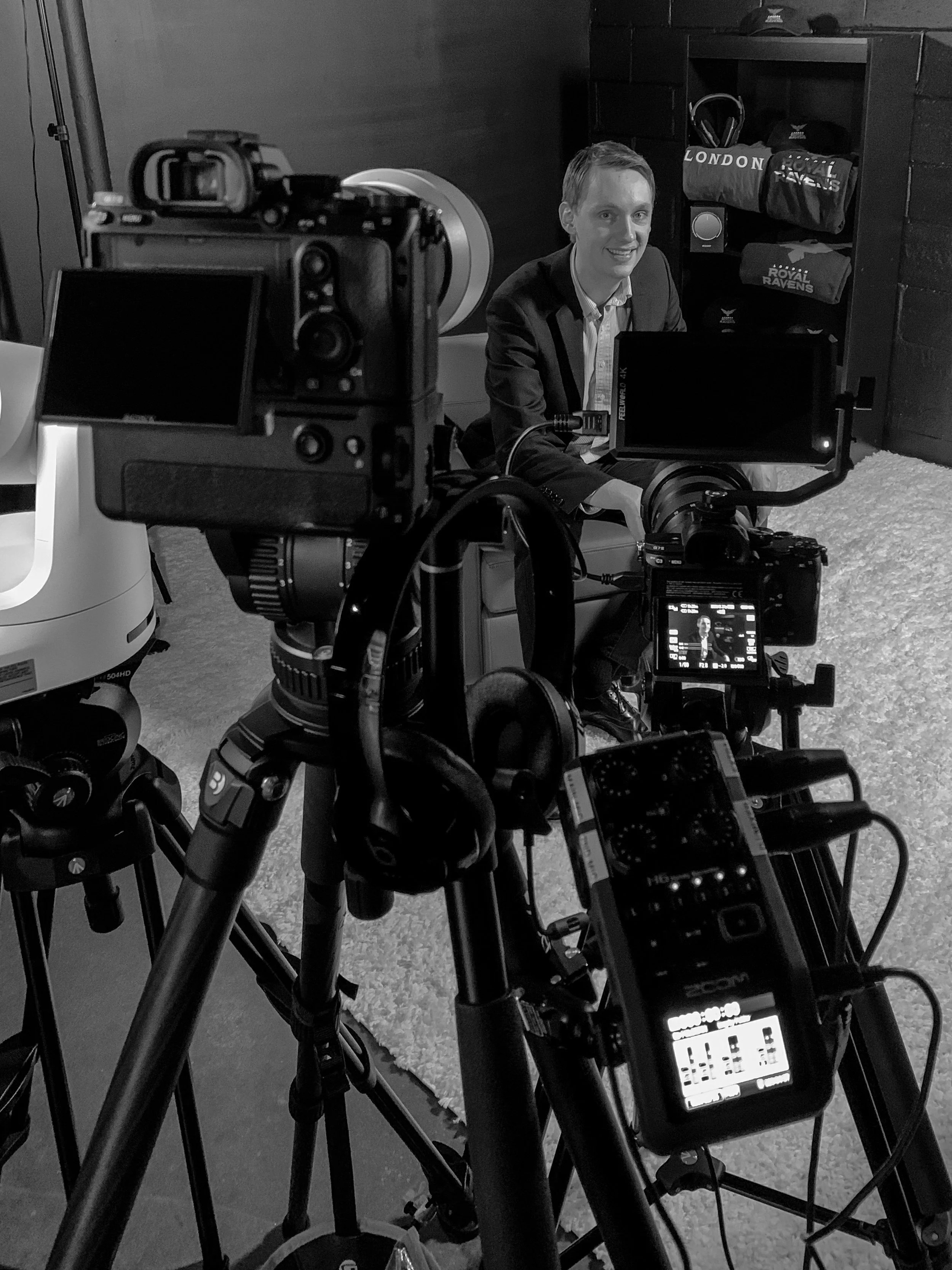 A man in a suit sitting on a chair, smiling, surrounded by professional cameras and equipment for filming or photography, with shelves holding bags labeled 'LONDON' and 'ROYAL RAVENS' in the background.