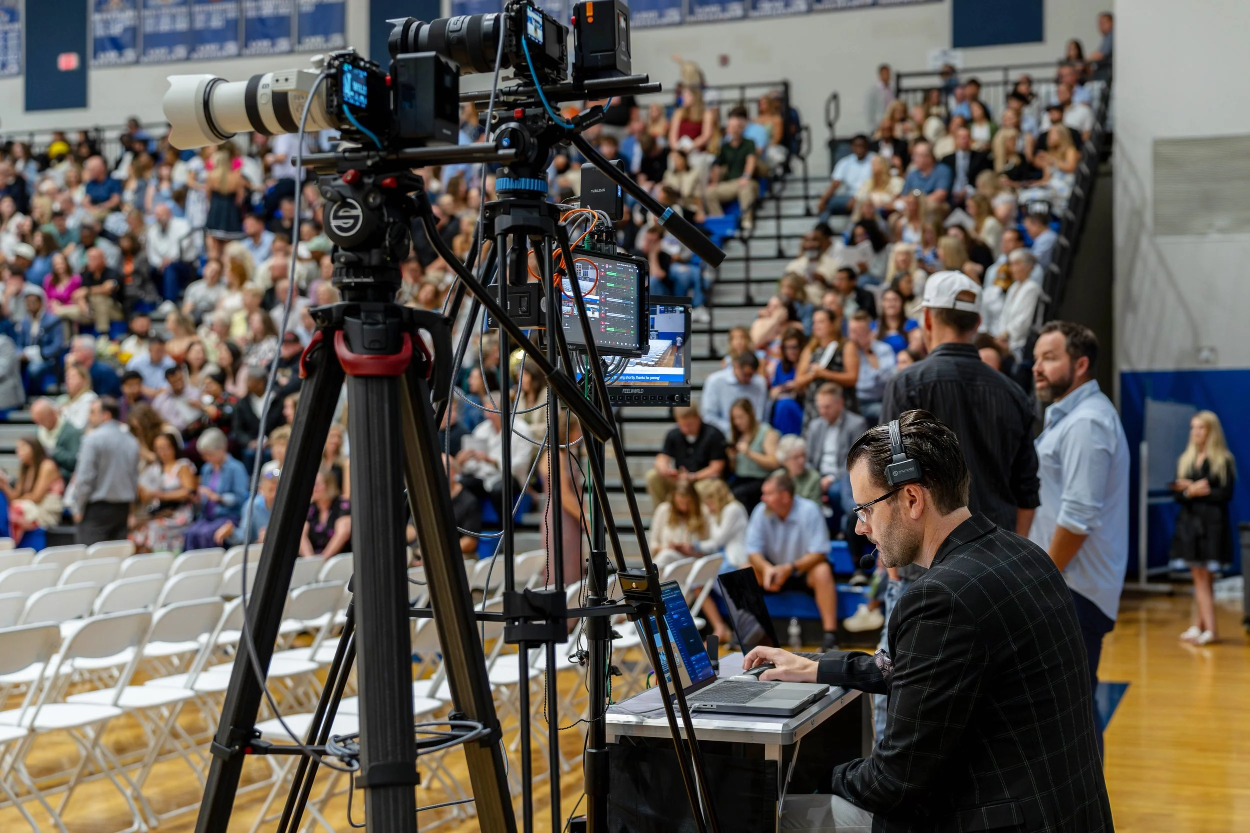 Professional video camera setup on tripod with a monitor, capturing an audience in a gymnasium during an event.