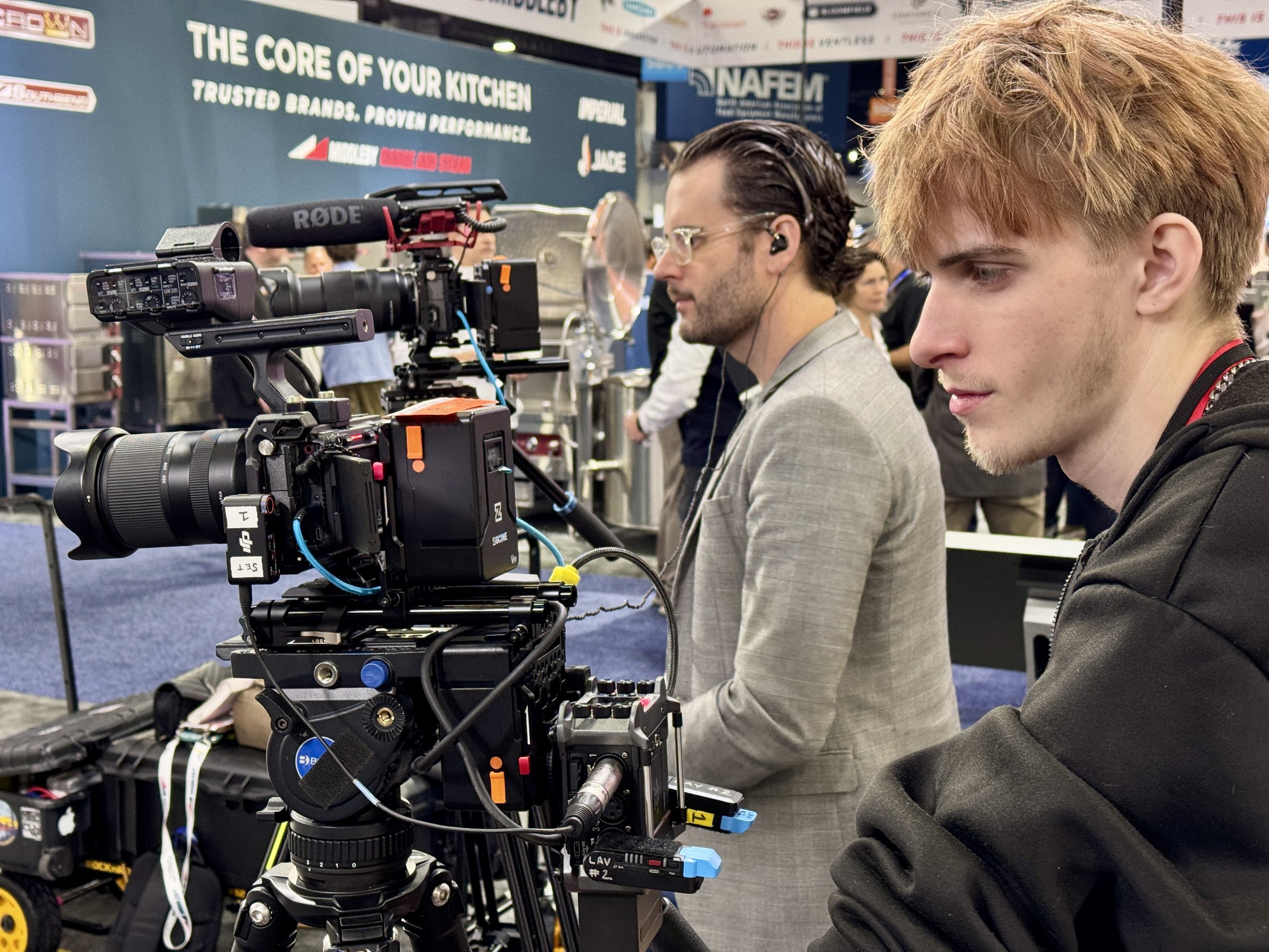 Two men operating professional video cameras at a trade show or event, with banners and equipment in the background.