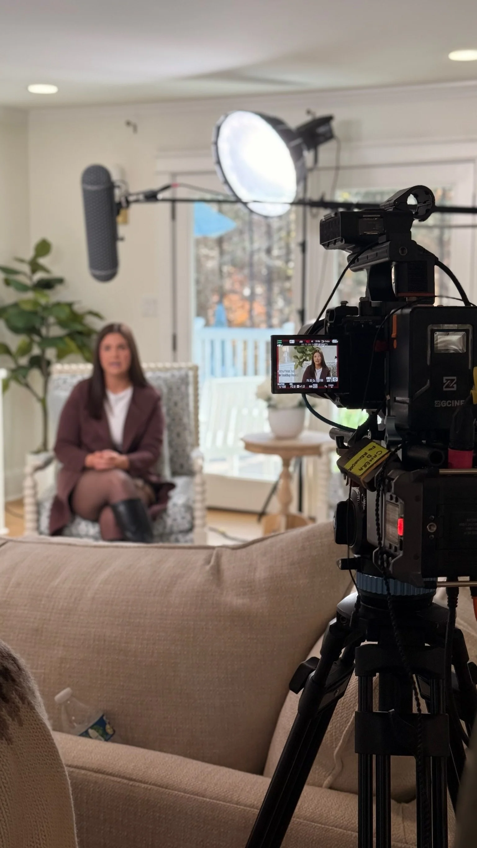 A woman being filmed during an interview, sitting on a patterned chair in a bright living room, with professional lighting, a background of windows and a sliding door leading to an outdoor space, and a large brown sofa in the foreground.