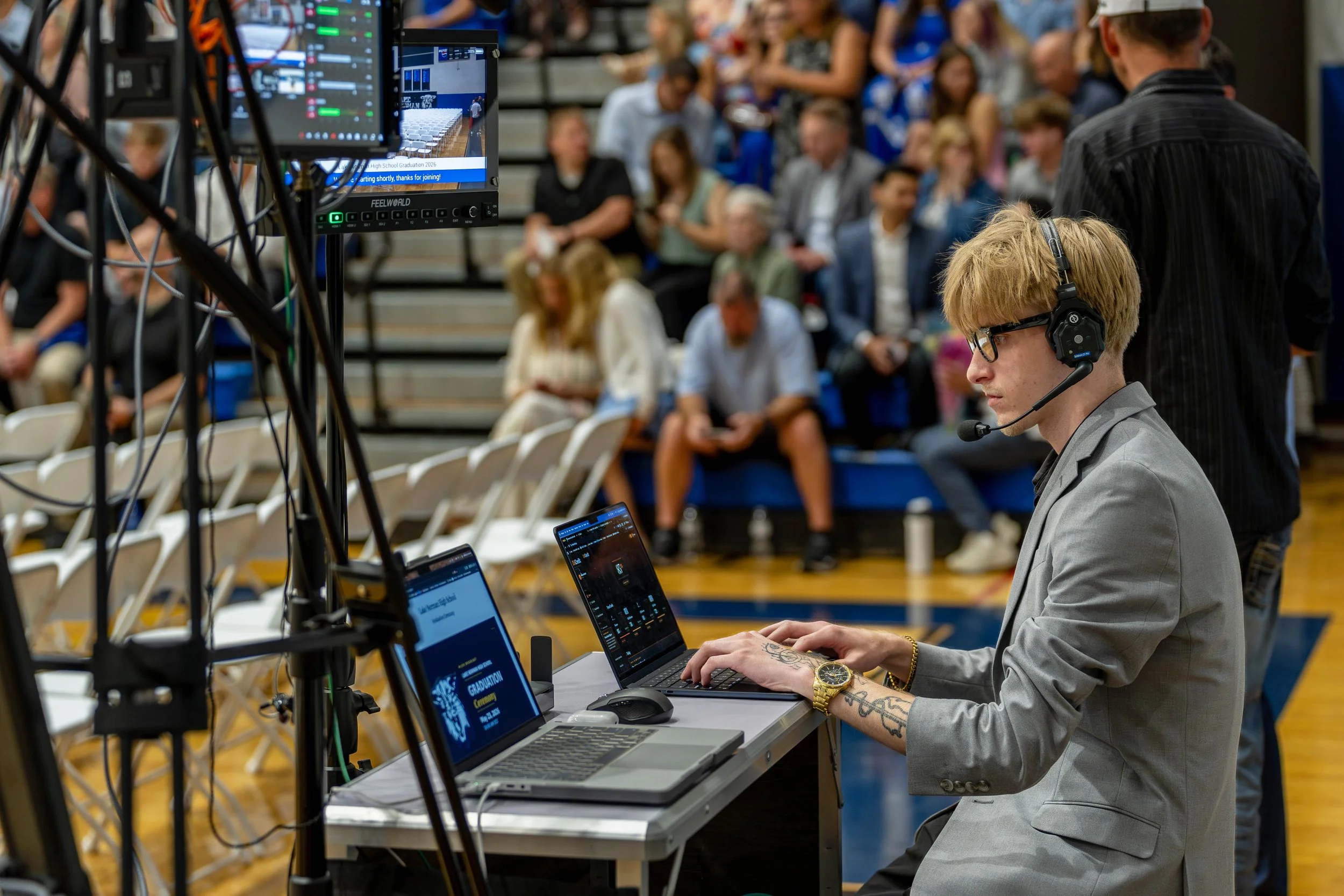A young man with blond hair, glasses, and tattoos on his arms, wearing a gray suit, is working on a laptop at a desk with multiple screens and audio equipment. In the background, there are many people seated in bleachers, attending an indoor event, and a monitor displays information about a high school graduation ceremony.