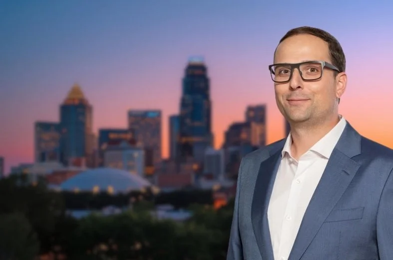 A man in a gray suit and white shirt standing outdoors with a city skyline in the background at sunset.