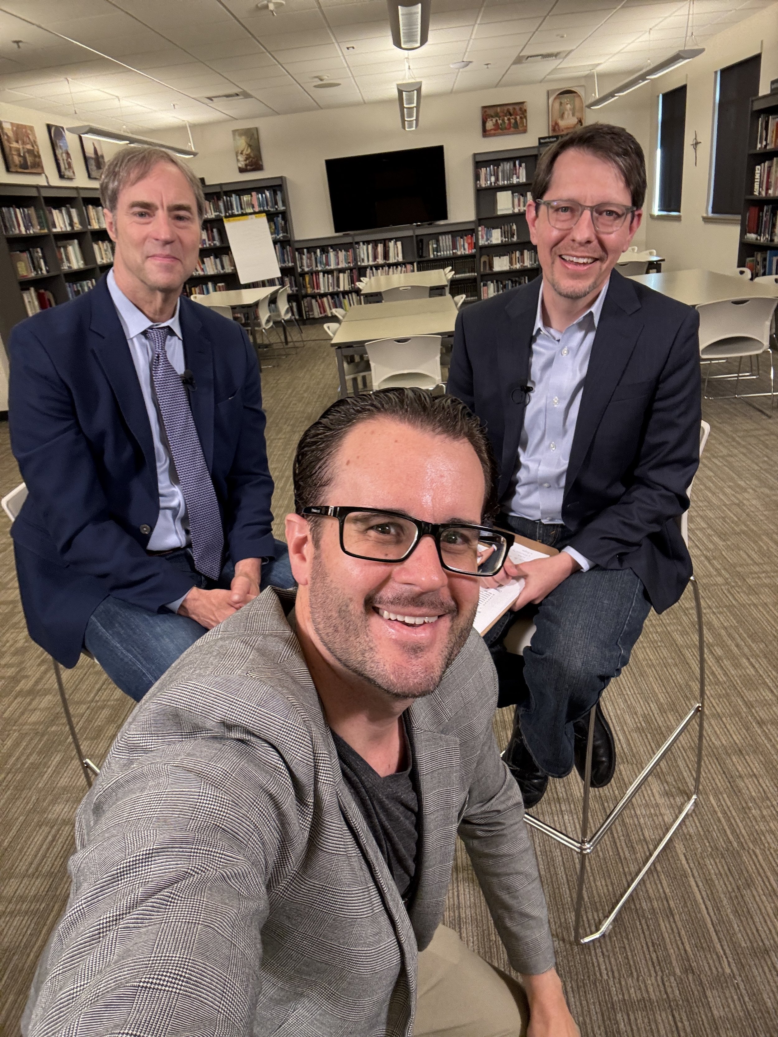 Three men are posing for a selfie in a library. The man in the front is wearing glasses and a grey plaid blazer, smiling at the camera. The two men behind him are seated at a table, both wearing dark suits with light shirts, smiling. The library has bookshelves, tables, chairs, and a large TV on the wall.