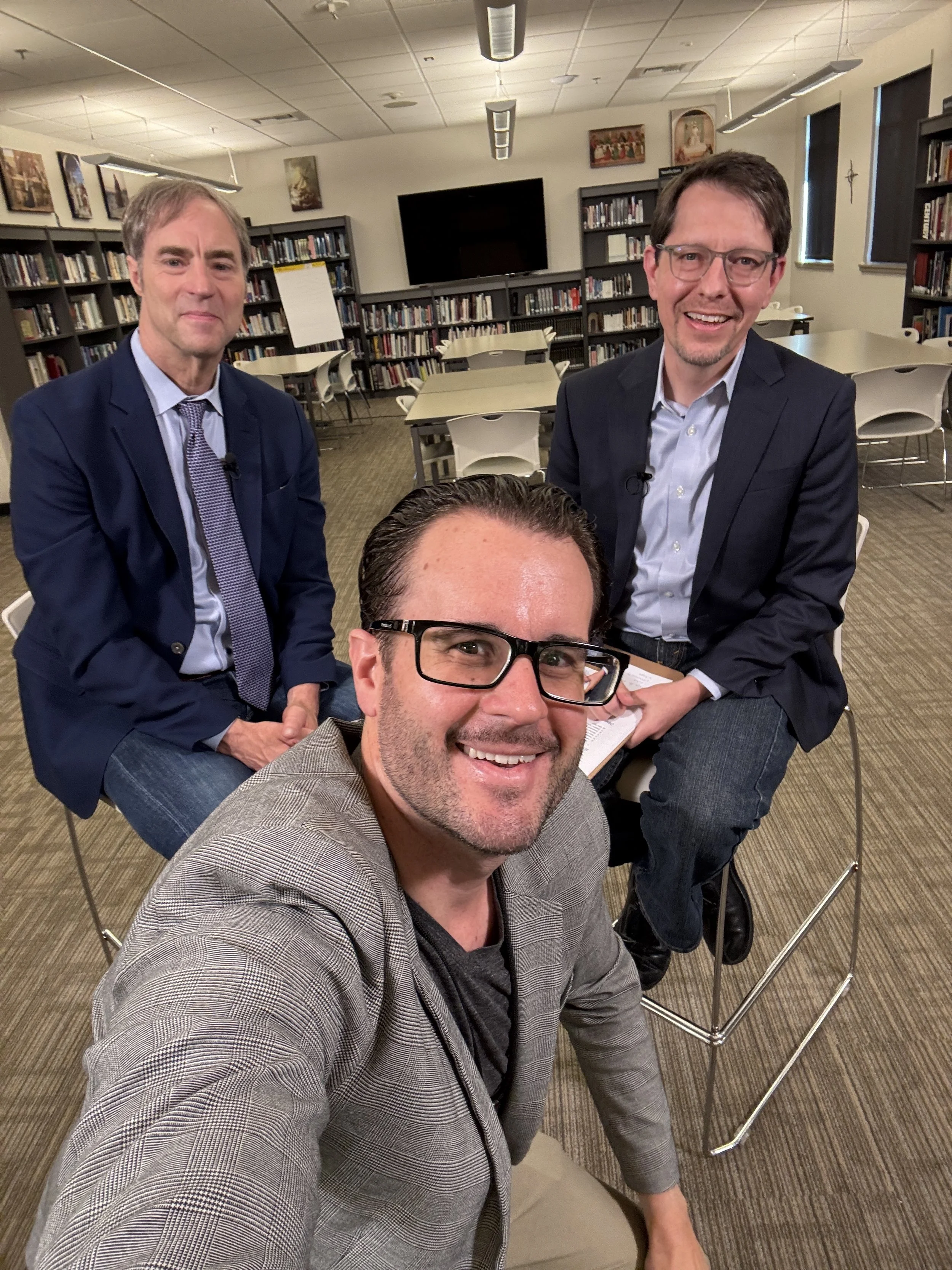 Three men are posing for a selfie in a library. The man in the front is wearing glasses and a grey plaid blazer, smiling at the camera. The two men behind him are seated at a table, both wearing dark suits with light shirts, smiling. The library has bookshelves, tables, chairs, and a large TV on the wall.