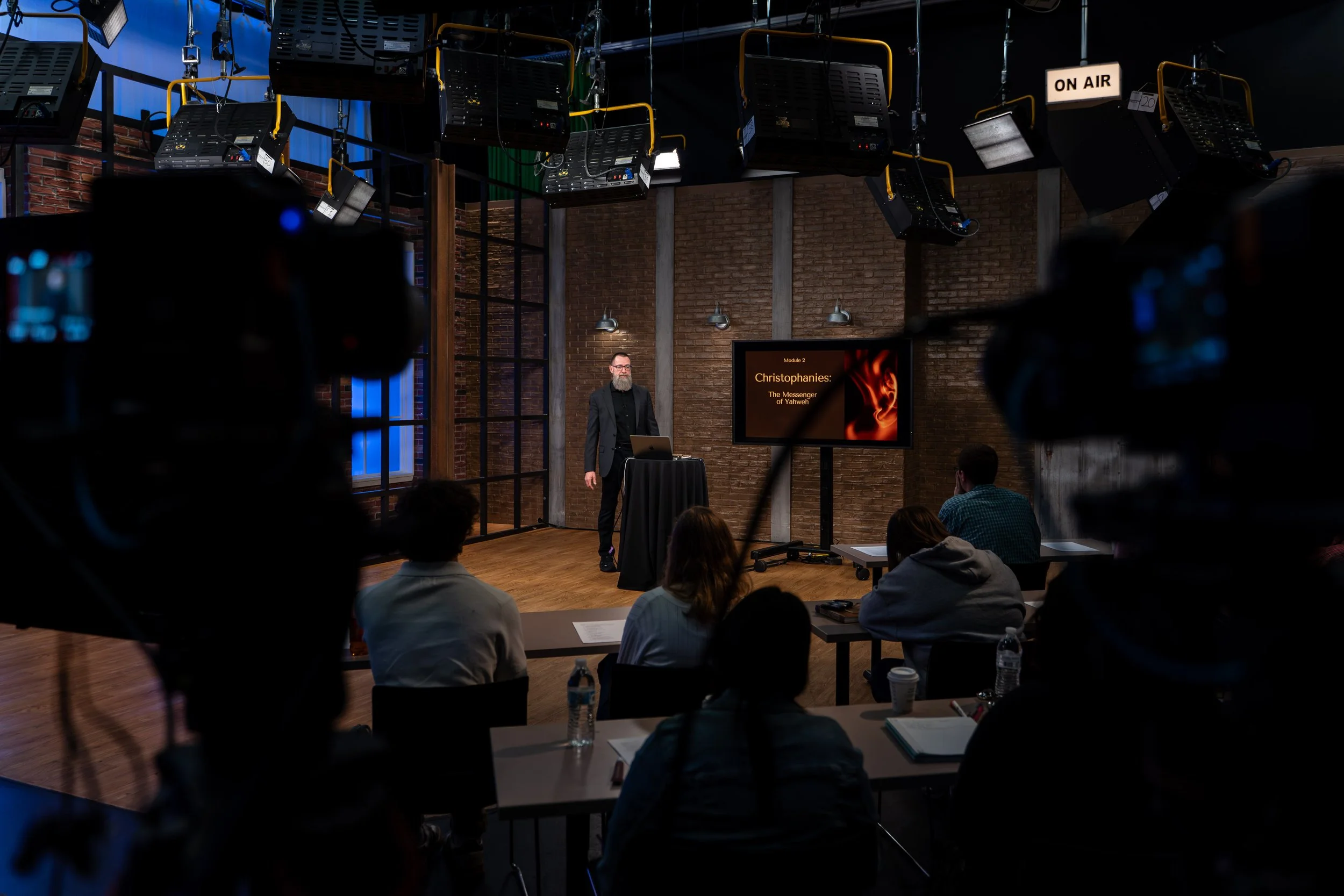 A presenter giving a lecture or presentation on a stage with a screen displaying the title 'Christophanies: The Messenger of Yahrweh' in a studio with lighting equipment overhead and an audience seated at tables.