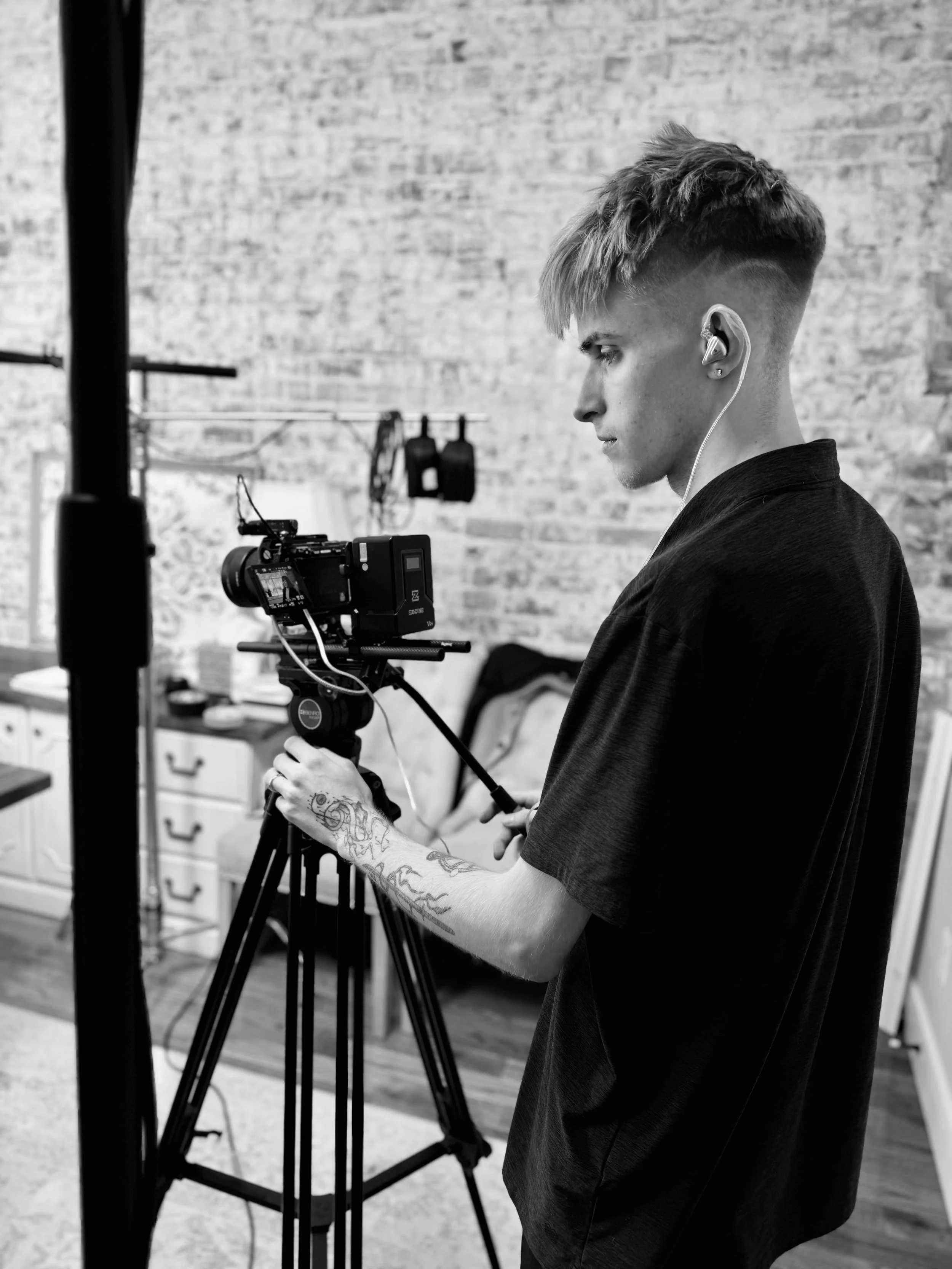 A young man operating camera equipment in a room with an exposed brick wall, wearing earphones, and focusing on the camera.