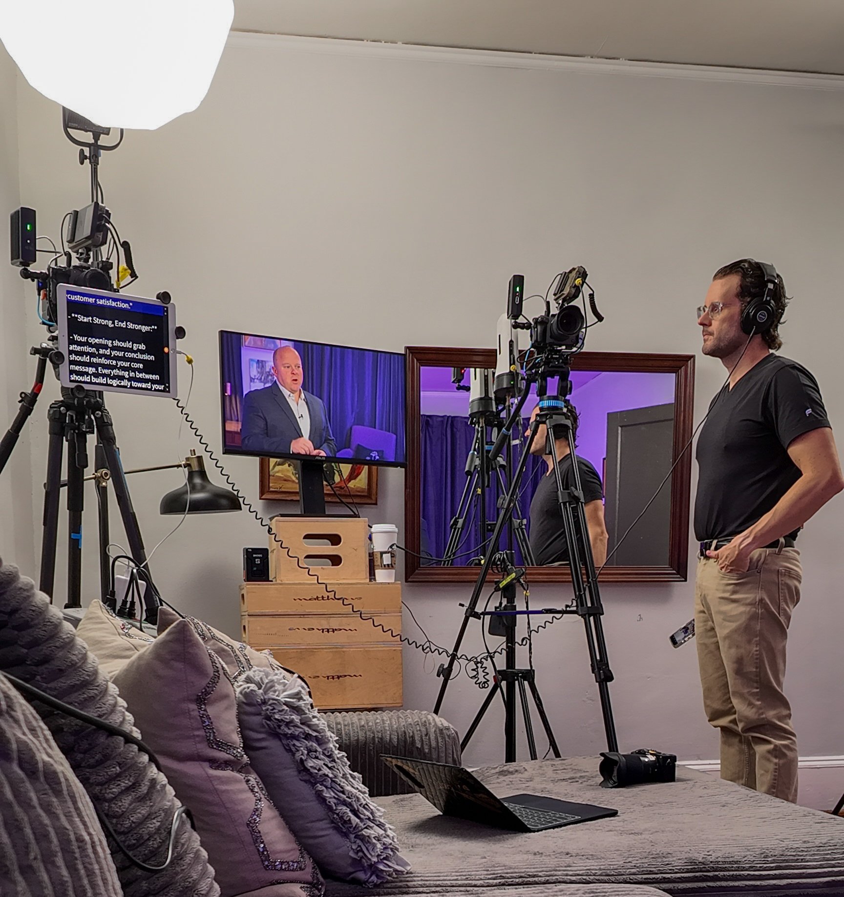 A behind-the-scenes look at a video recording setup with a man in black shirt and beige pants operating cameras, large studio lights, a monitor showing a speaker, and a mirror reflecting the scene.
