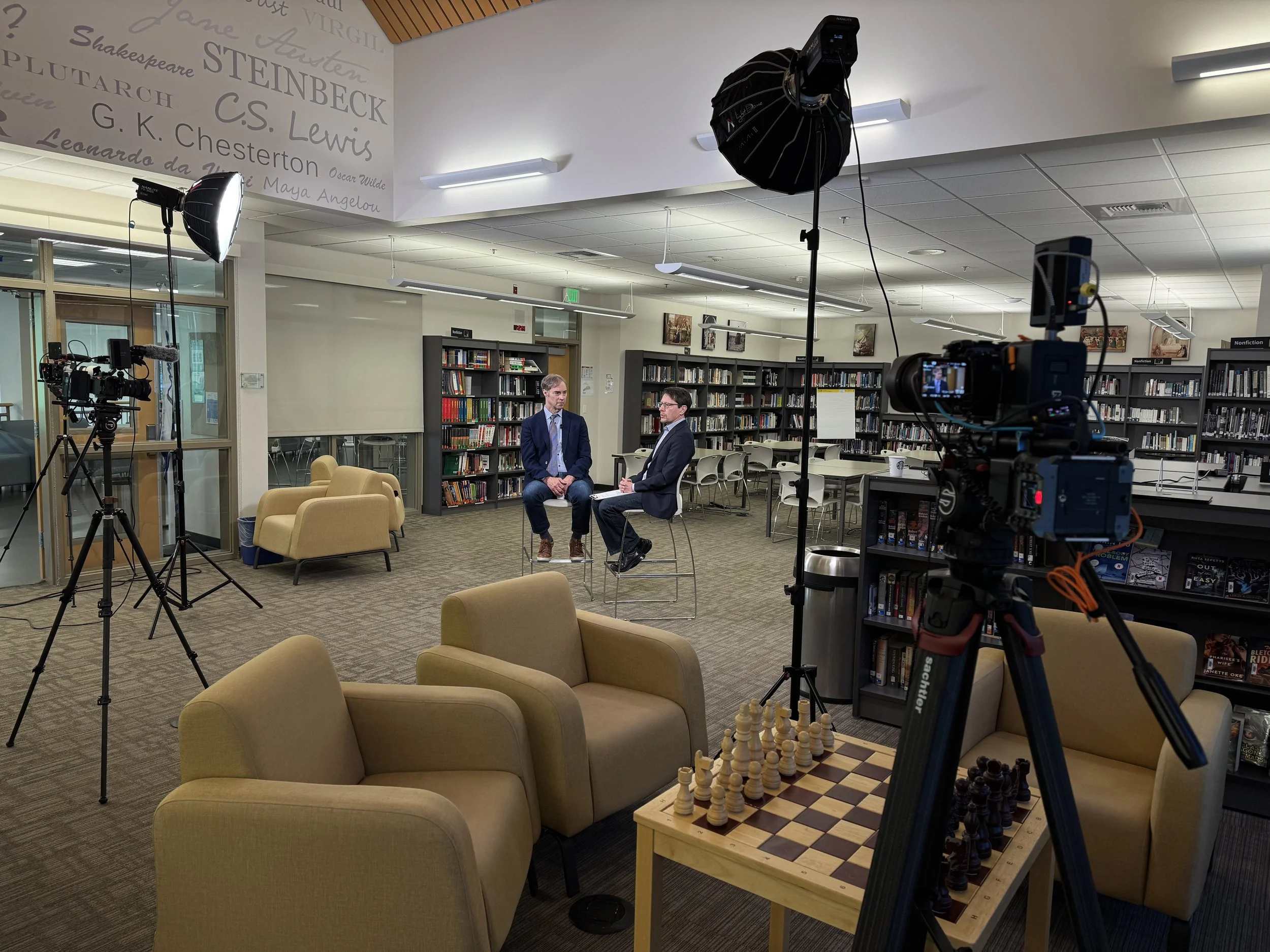 Two men in suits being interviewed in a library surrounded by furniture, bookshelves, cameras, and lighting equipment for recording.