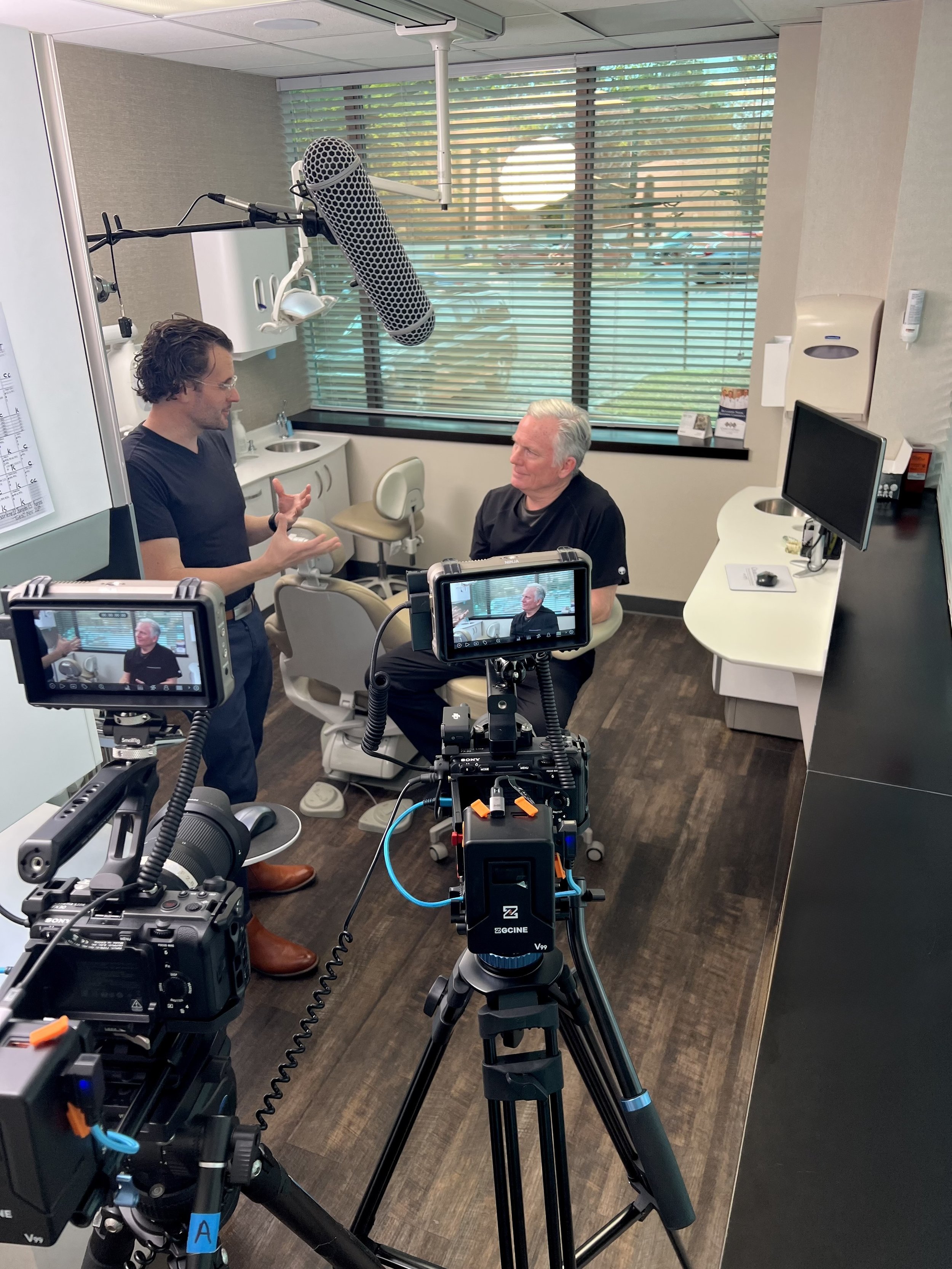 A doctor and a patient in a clinical office are recording a conversation using professional video equipment. The doctor is standing and speaking, while the patient is seated and listening. The room has a large window with blinds, a computer monitor, and medical equipment.