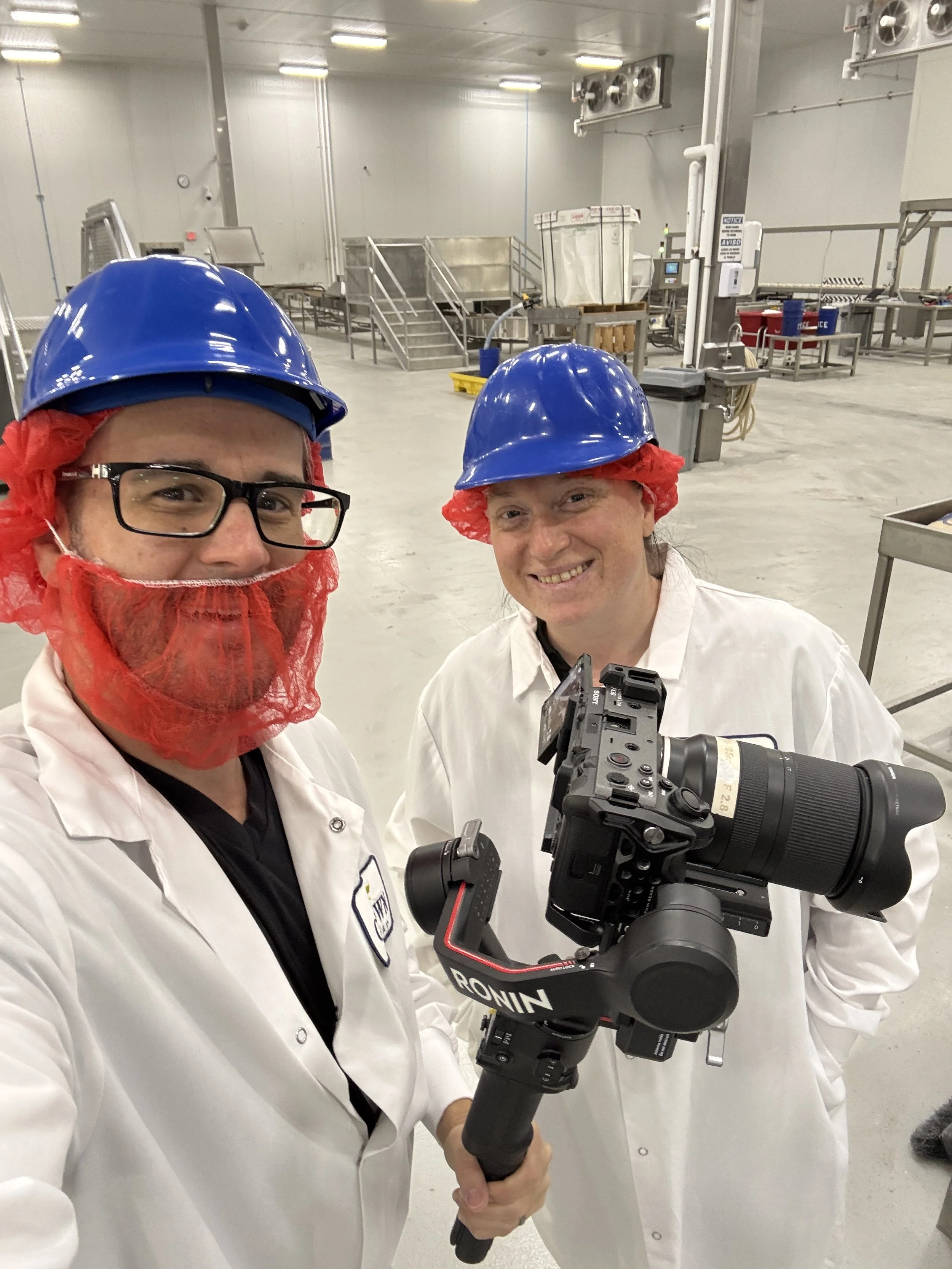 Two people wearing blue safety helmets, red hair nets, white lab coats, and glasses smiling for a photo in an industrial or food processing facility. One person is holding a camera mounted on a stabilizer.