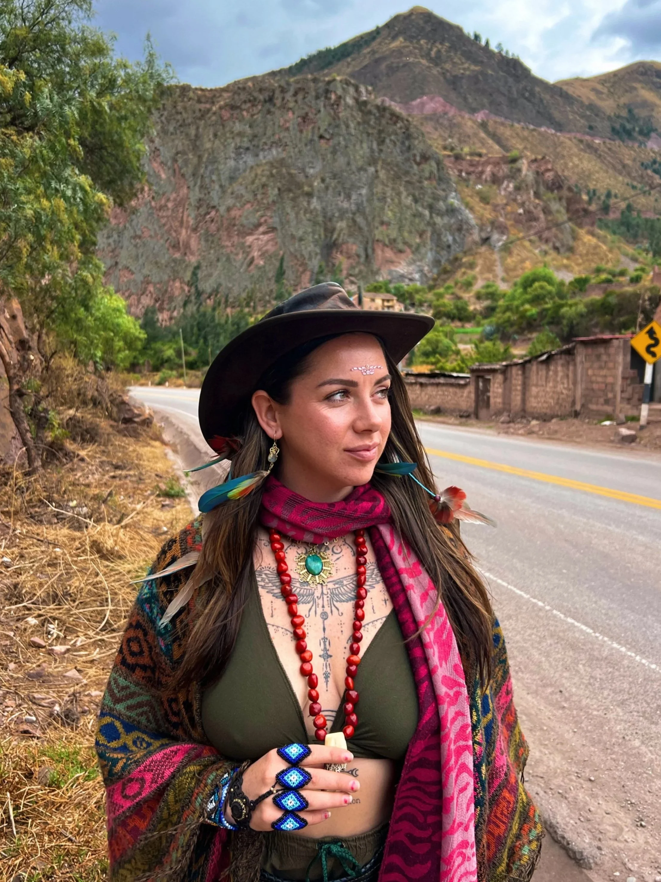 A woman standing by a roadside in a mountainous area, wearing a wide-brimmed hat, colorful accessories, jewelry, and a shawl, with mountains and a cloudy sky in the background.