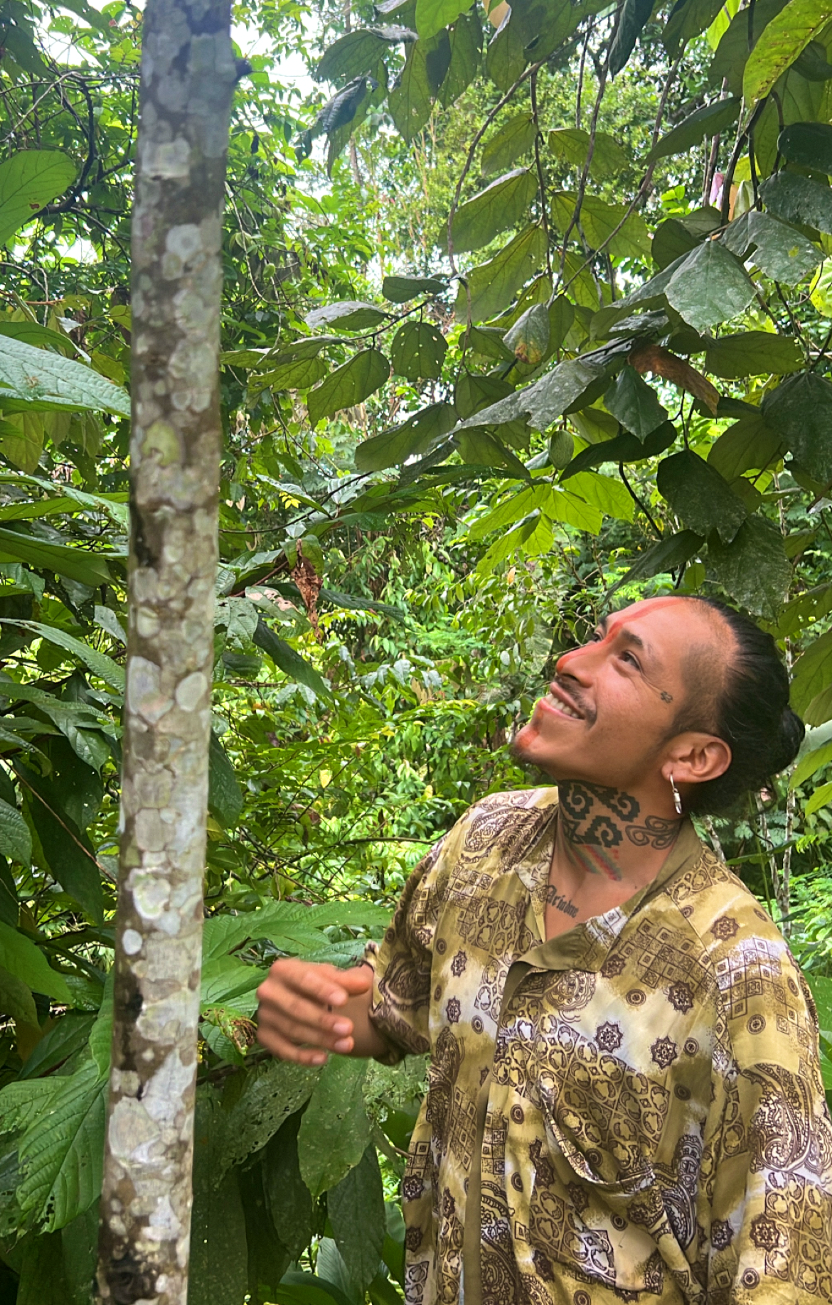 A person with dark hair pulled back, colorful tattoos on their neck and chest, wearing a patterned shirt, smiling and looking up in a lush, green jungle environment.
