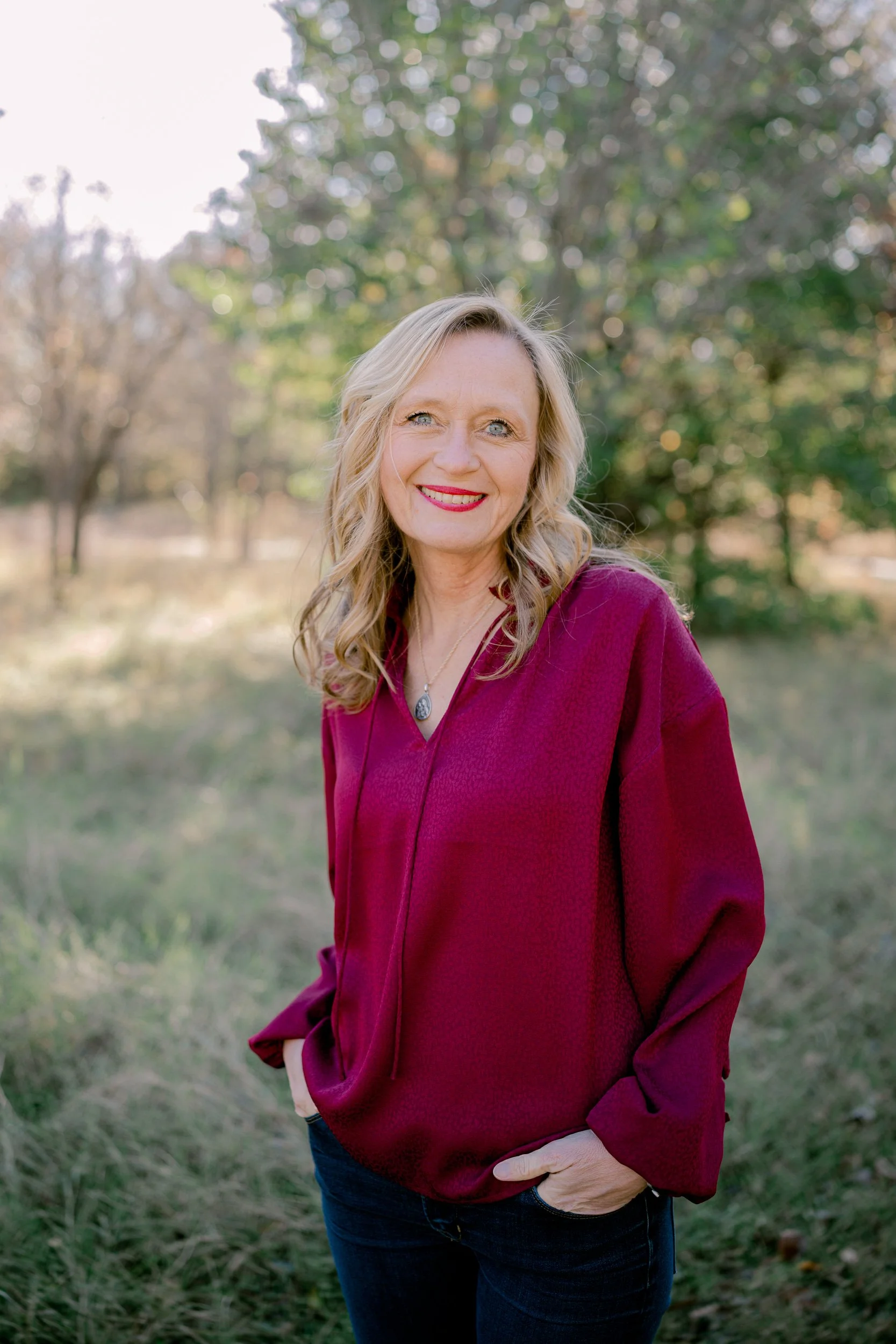 A woman with blonde hair, wearing a burgundy top and jeans, smiling outdoors with trees in the background.
