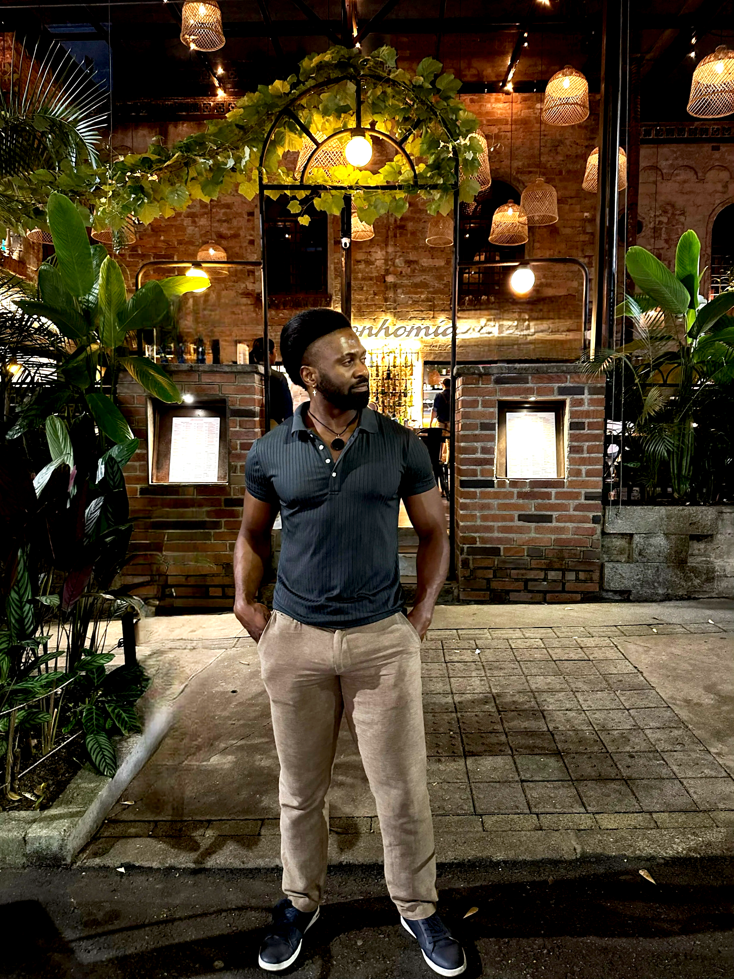 A man standing outside a restaurant or cafe at night, with hands in his pockets, wearing a black striped polo shirt, beige pants, and sneakers, with greenery around and a brick building behind him.
