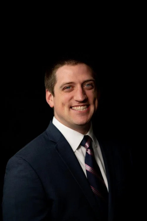 A smiling young man in a dark suit, white shirt, and striped tie posing against a black background.