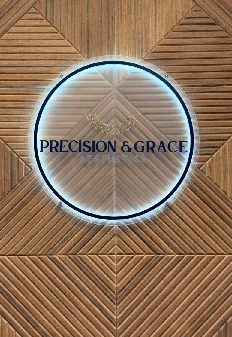 A round illuminated sign with the words 'Precision & Grace Aesthetics' on a wooden textured wall.
