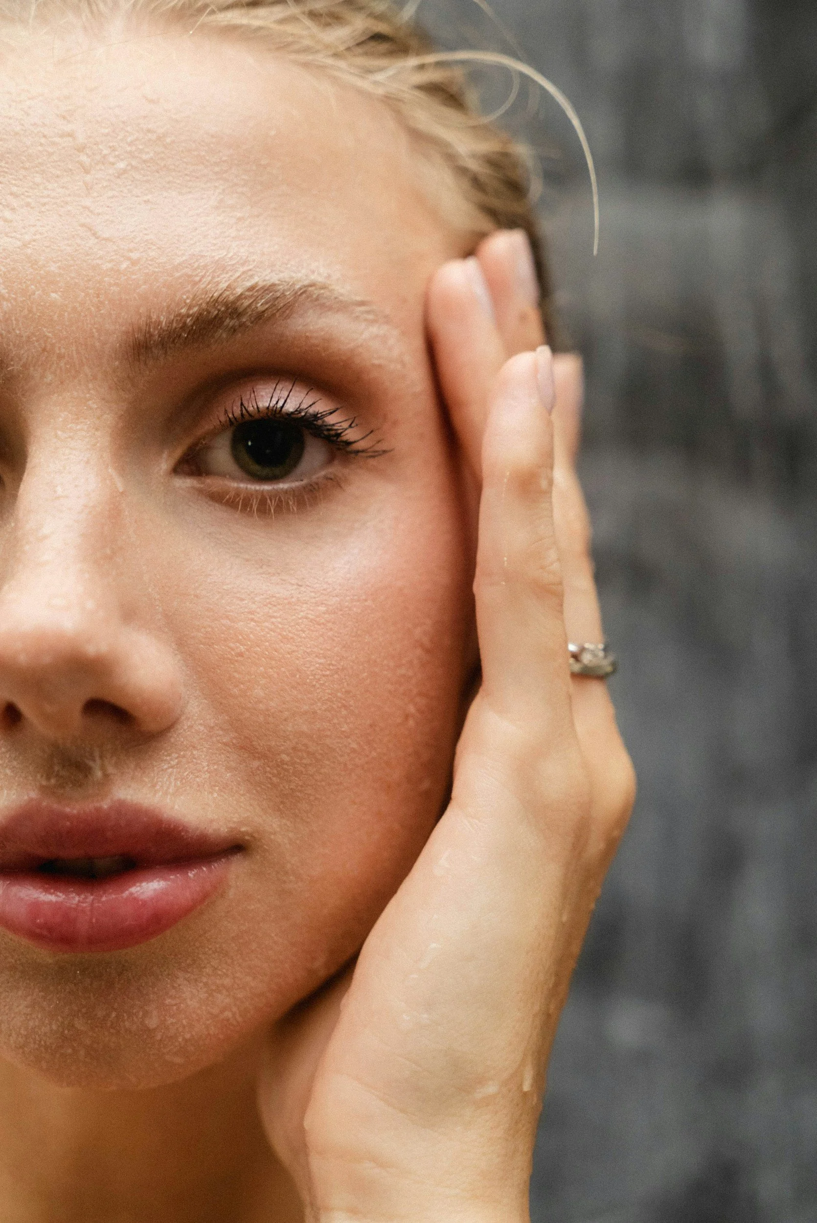 A close-up of a woman with fair skin, green eyes, and blonde hair, touching her face with her hand, wearing a wedding ring.