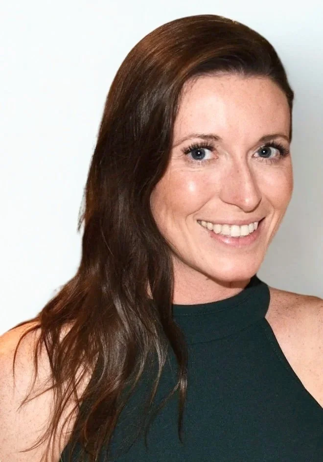 A woman with long brown hair, blue eyes, and fair skin, smiling at the camera, wearing a dark sleeveless top.