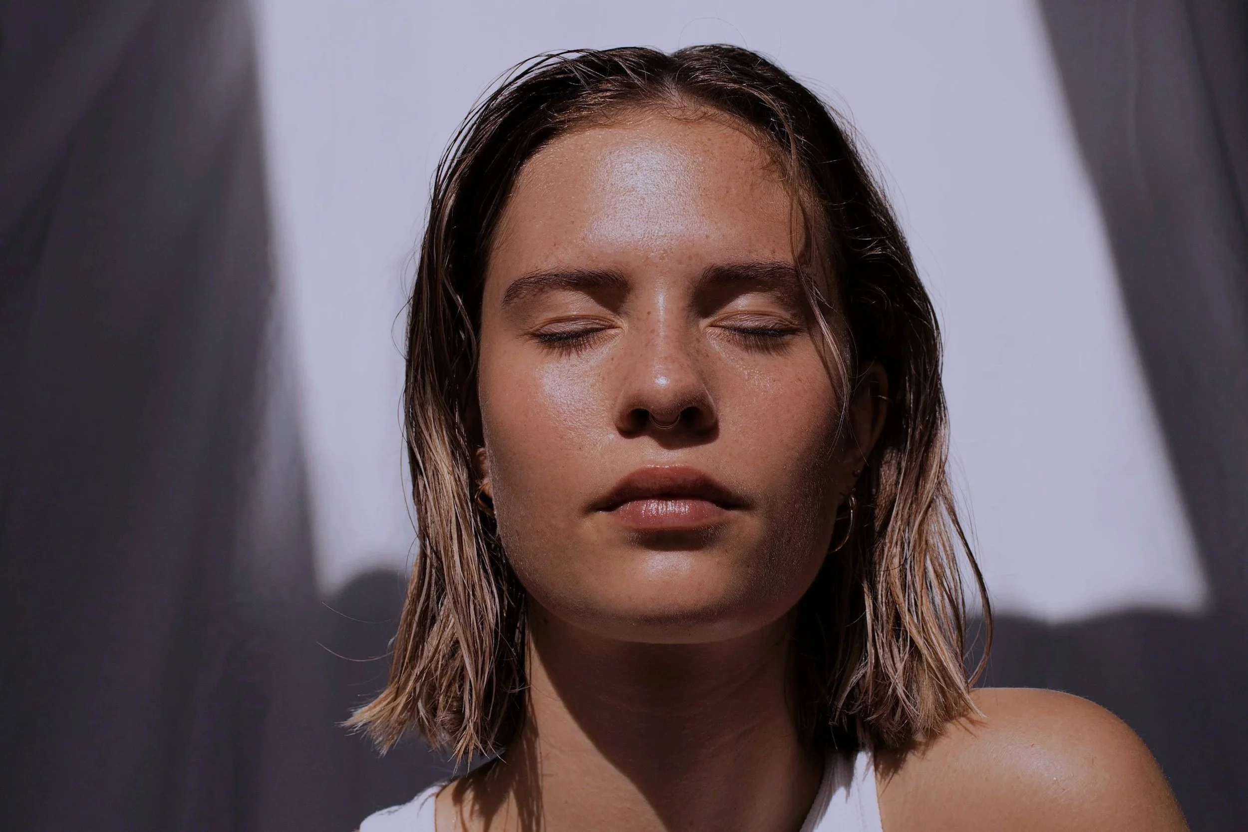 A young woman with wet hair and closed eyes, in front of a white background with dark shadows.