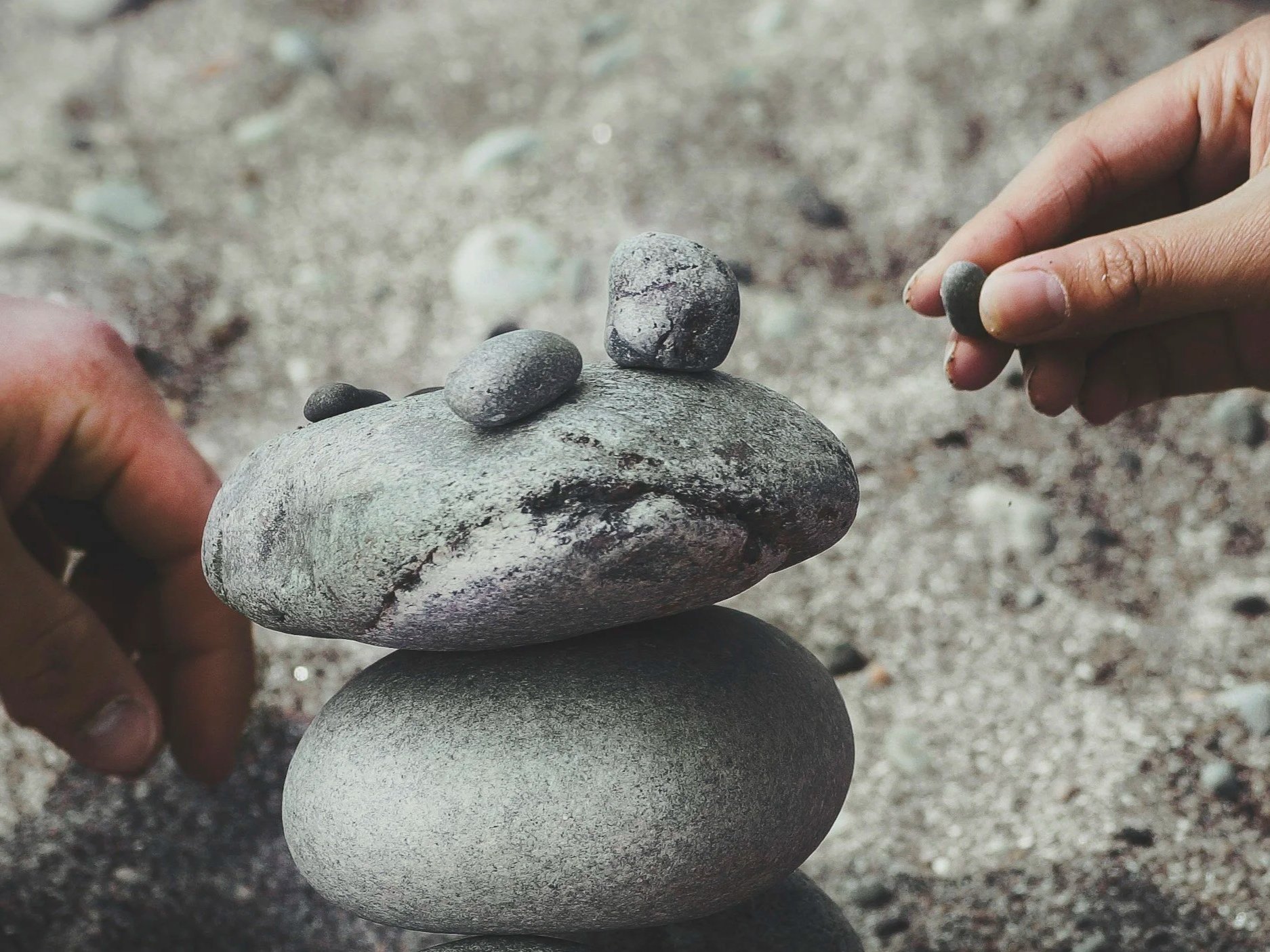 Person balancing three small rocks on top of two larger rocks on a sandy ground.