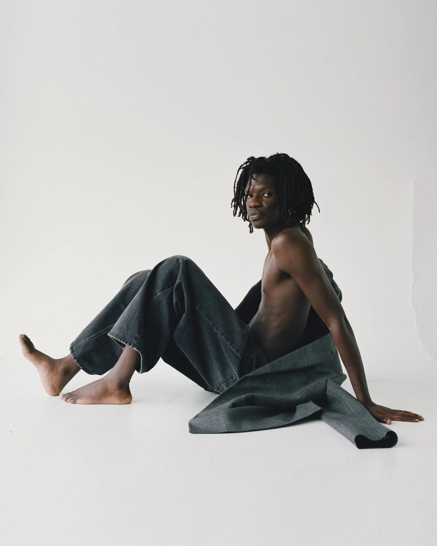 Image of a black male model sitting profile on the floor of a white studio wearing large black jeans with a grey blazer draped around his body