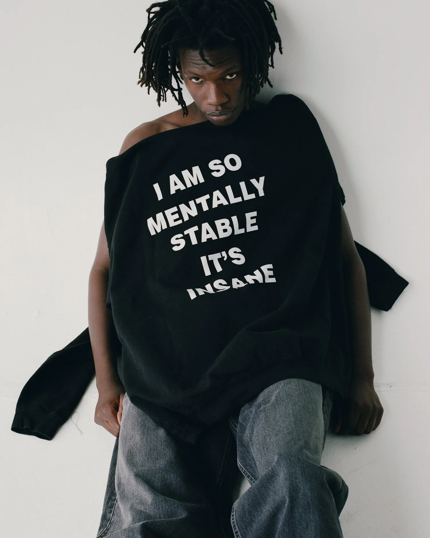 Portrait of a black male model laying down in a white studio with a hoodie that reads "I'm so mentally stable it's insane" on top of him