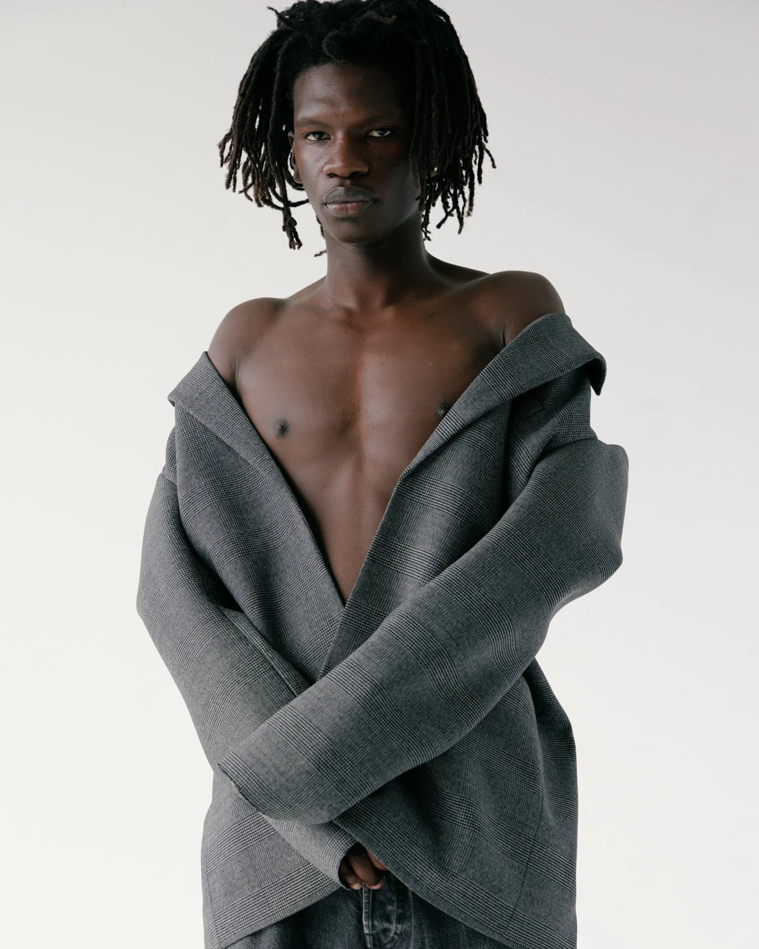 Image of a black male model with micro dreads bare chested wearing a grey blazer in a white studio