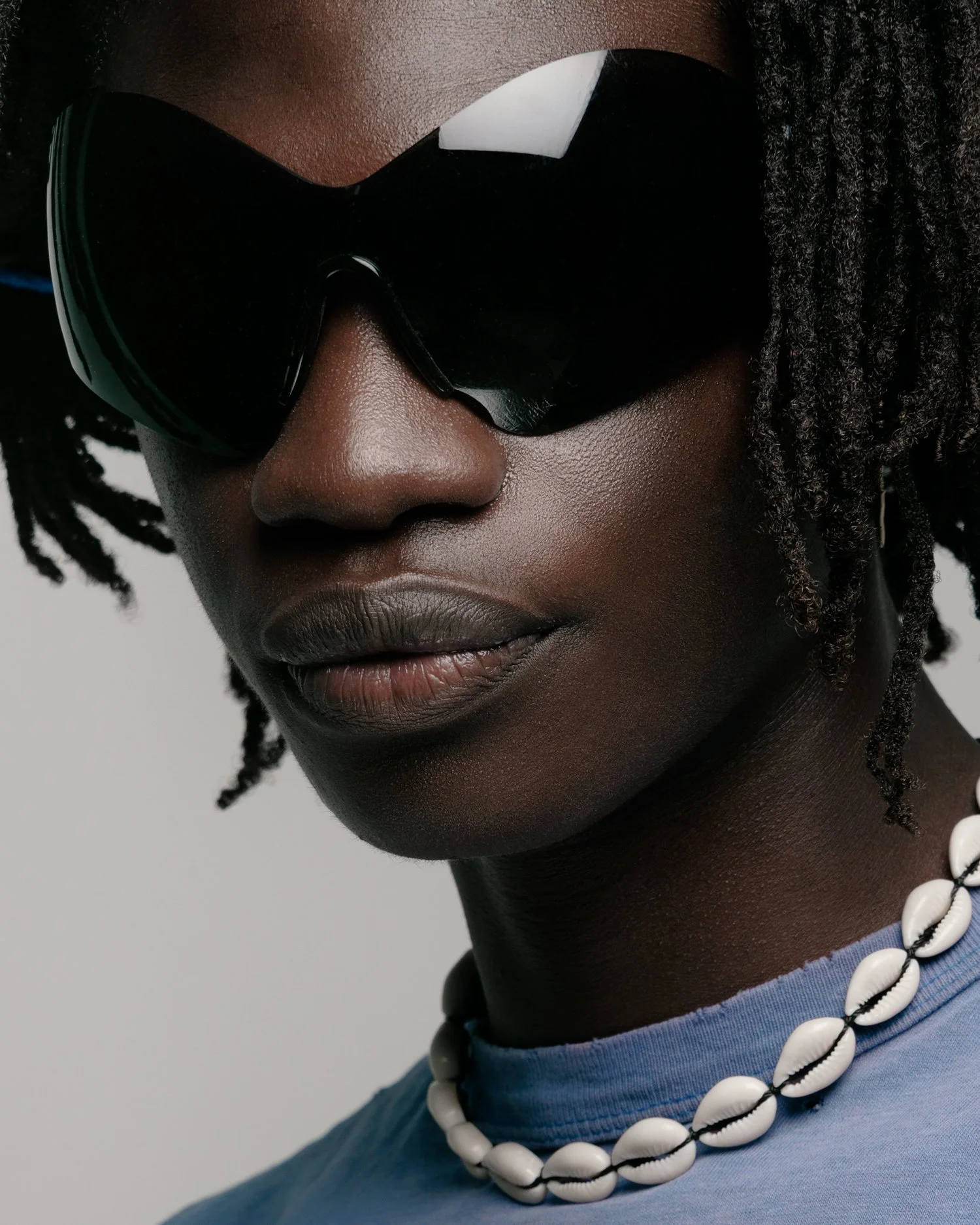 Portrait of a black male model with micro dreads wearing large black sunglasses, a torn sports tee, and a shell necklace