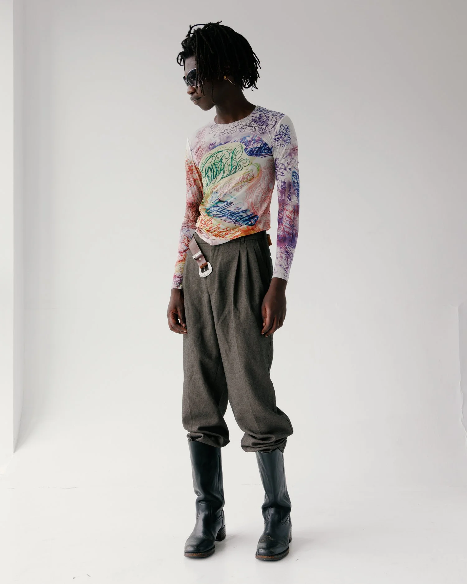 Image of a black male model in a white studio wearing a faux tattoo shirt, brown pants, a brown belt, and tall black boots