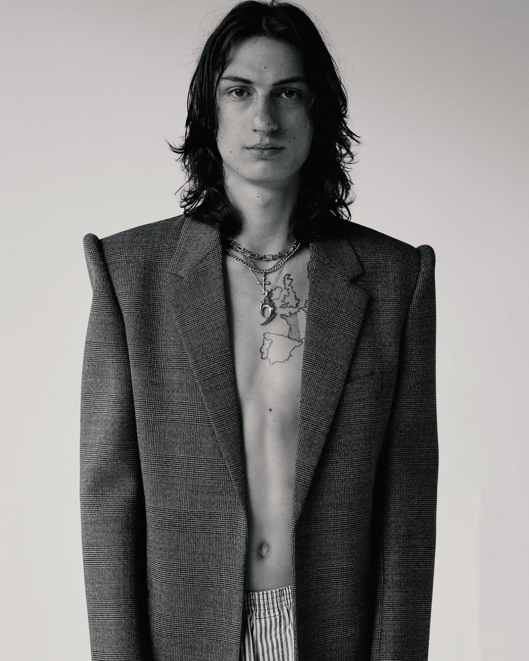 Black and white portrait of a white male model with long hair wearing a grey blazer, striped boxers, and silver necklaces in a white studio