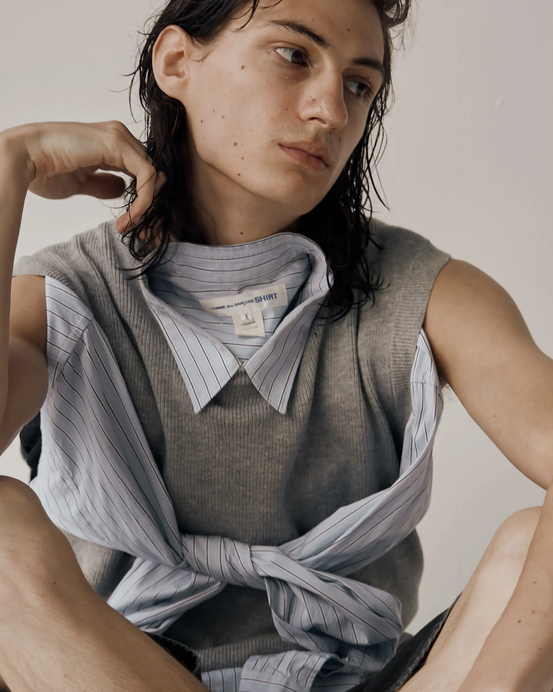 Portrait of a white male model with long hair sitting on the floor of a white studio wearing a grey vest with a pinstripe button down shirt tucked into it