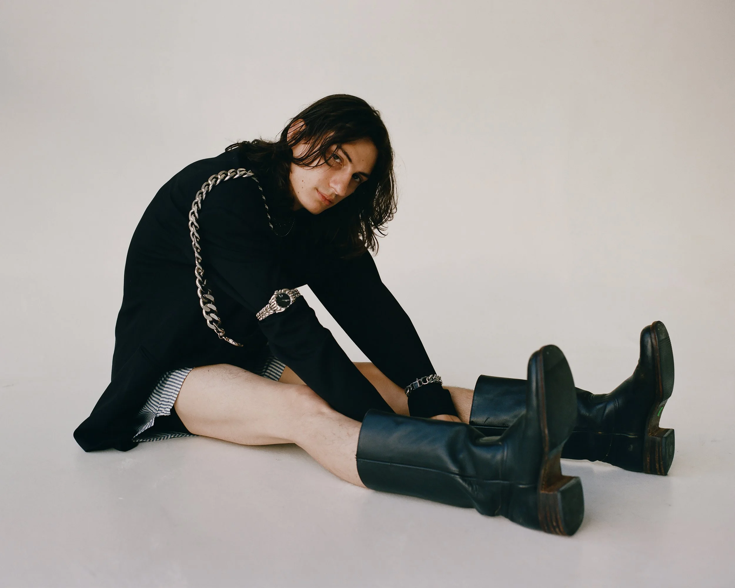 Image of a white male model with long hair sitting on the floor of a white studio wearing a black blazer, striped boxers, black boots, and silver jewelry