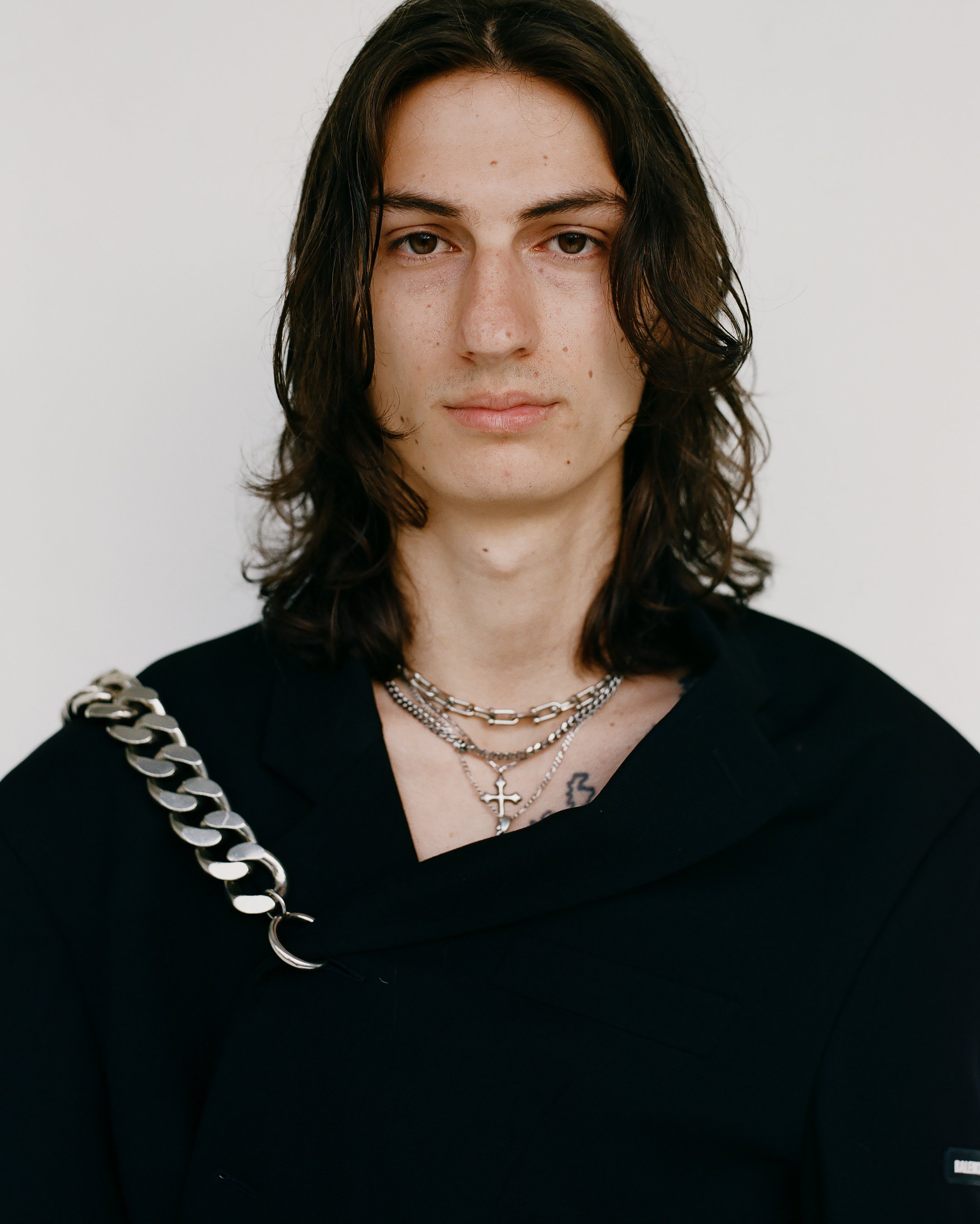 Portrait of a white male model with long brown hair wearing a black blazer and silver jewelry