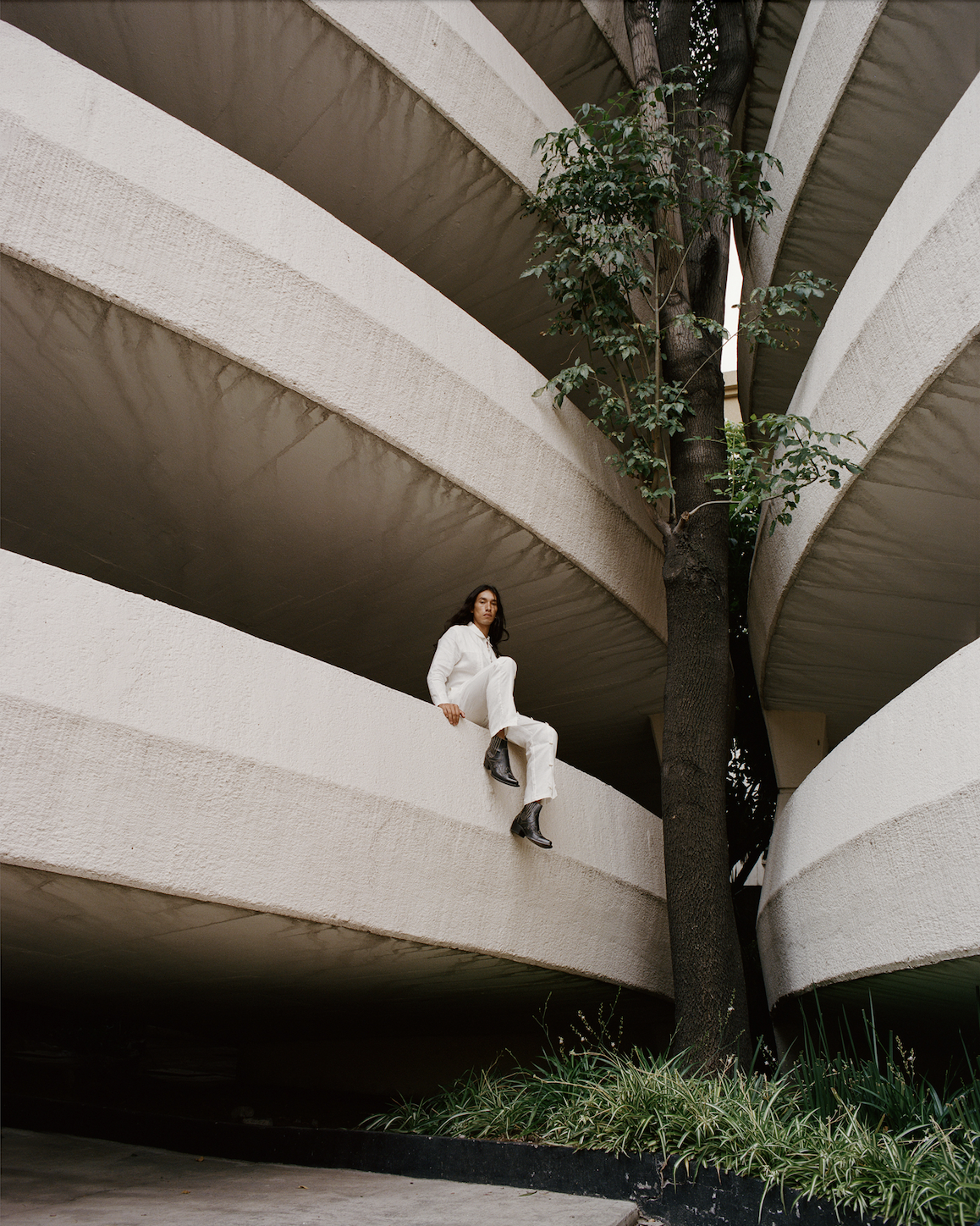 Image of a Mexican male model in a white outfit and cowboy boots sitting on a spiral parking garage ledge.