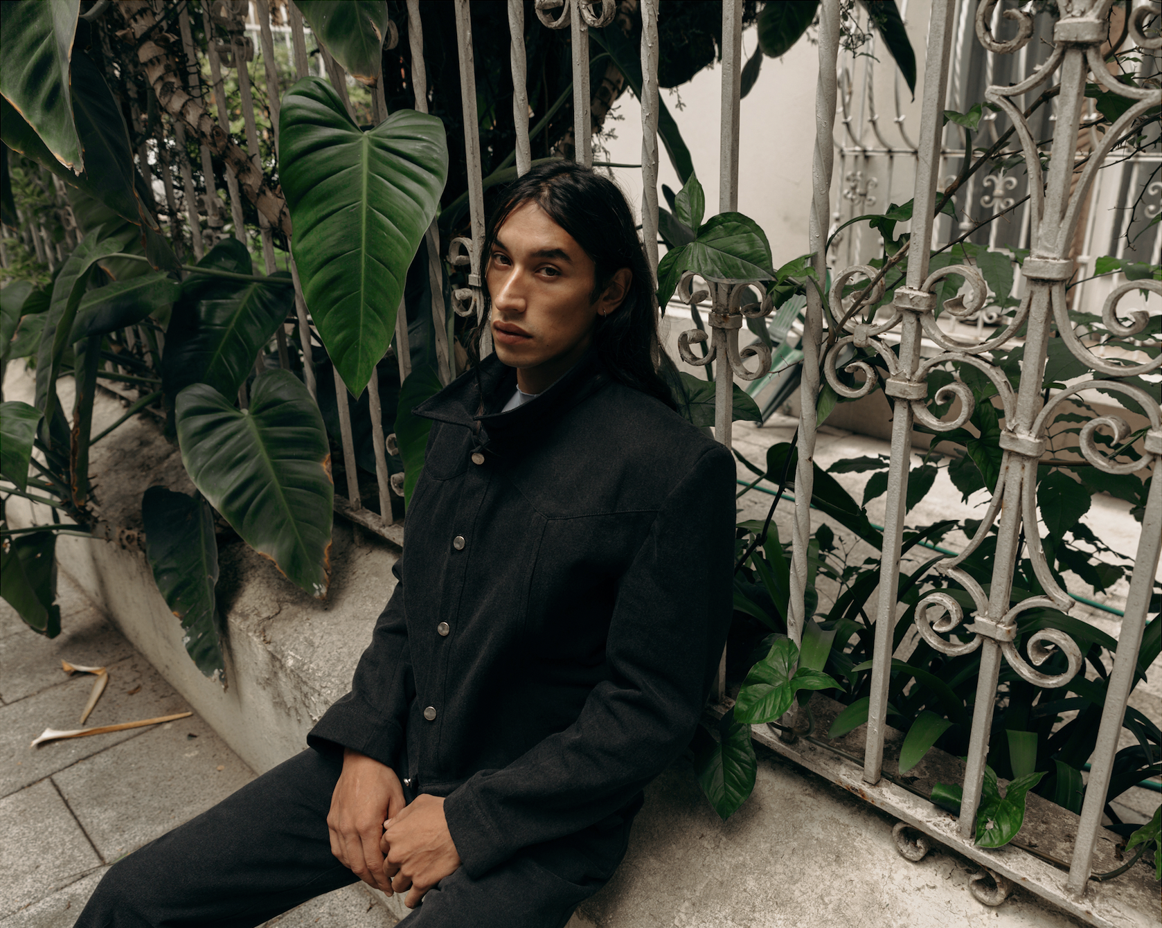 Image of a Mexican male model with long hair wearing dark grey clothes with lush green plants in the background