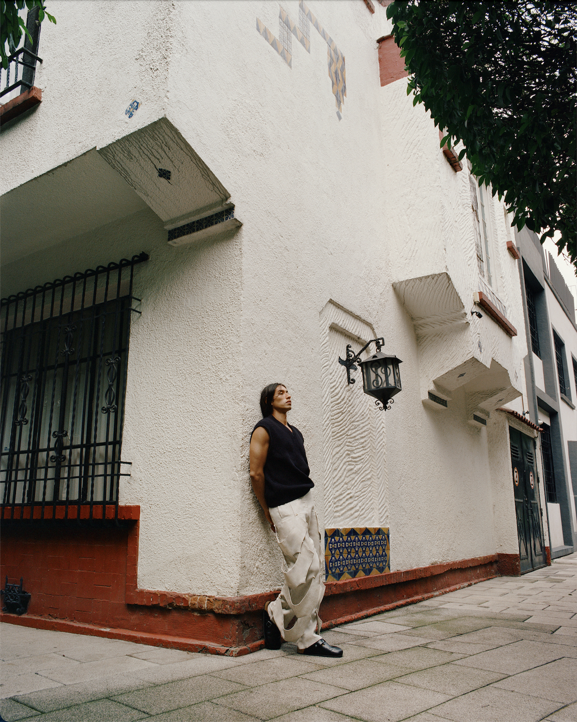 Image of a Mexican male model leaning on the wall of a building wearing a black vest and draped white trousers