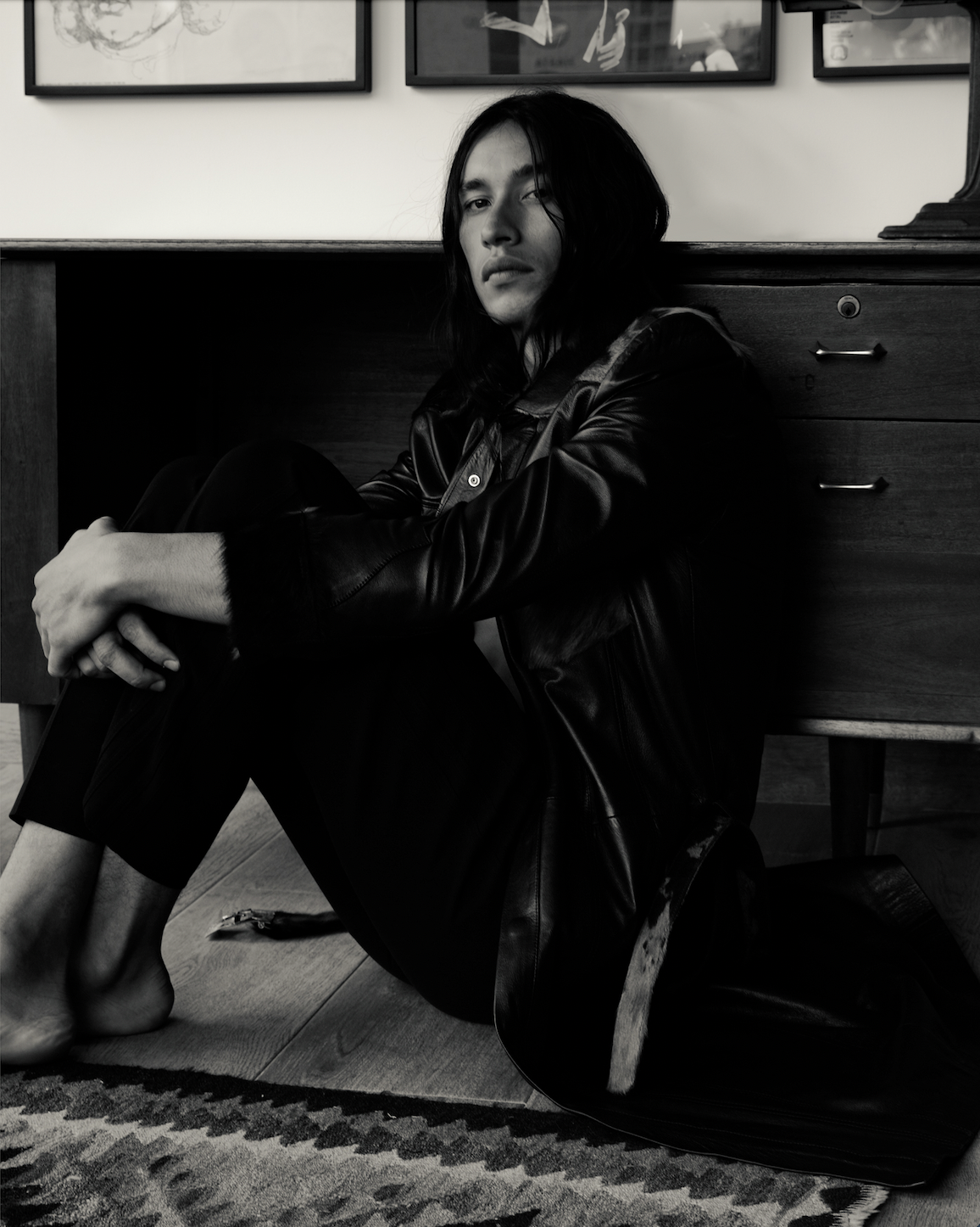 Black and white image of a Mexican male model sitting on the floor of an apartment wearing a black leather coat
