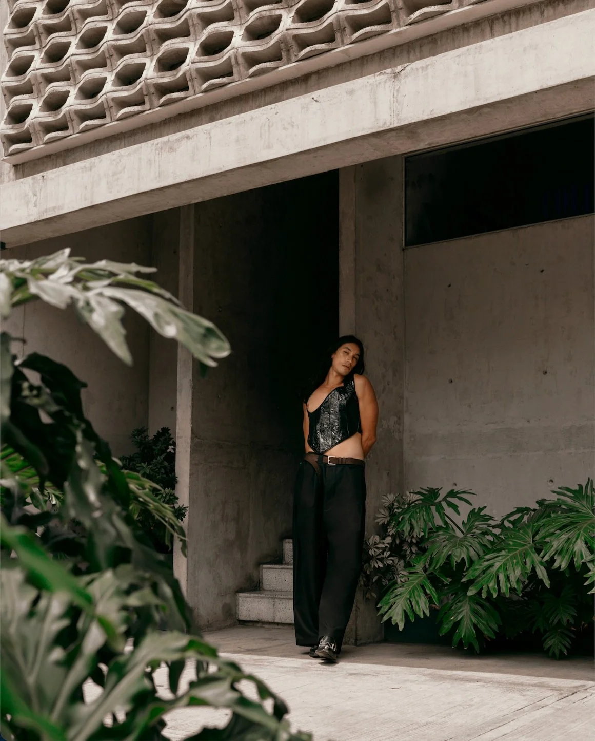 Image of a Mexican male model under the overhang of an apartment building wearing large black trousers and a shiny black corset top with plants surrounding him