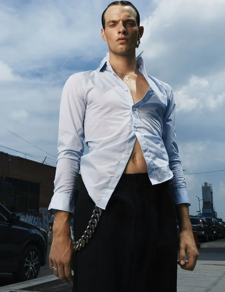 image of a white male model wearing a tight blue button down shirt, black trousers, and a silver oversize wallet chain on the street