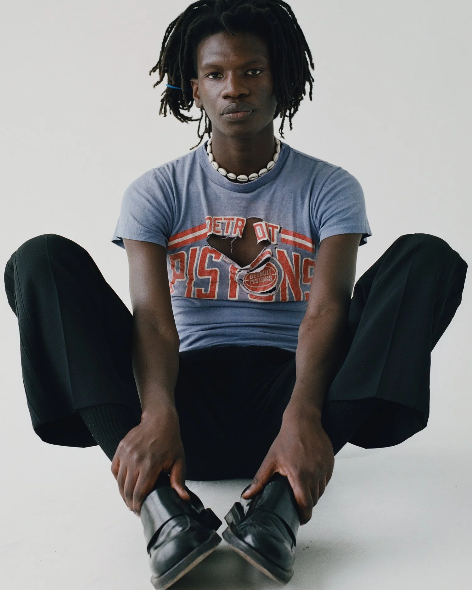 image of a black male model wearing a ripped sports t-shirt, black trousers and loafers, and a shell necklace in a white studio